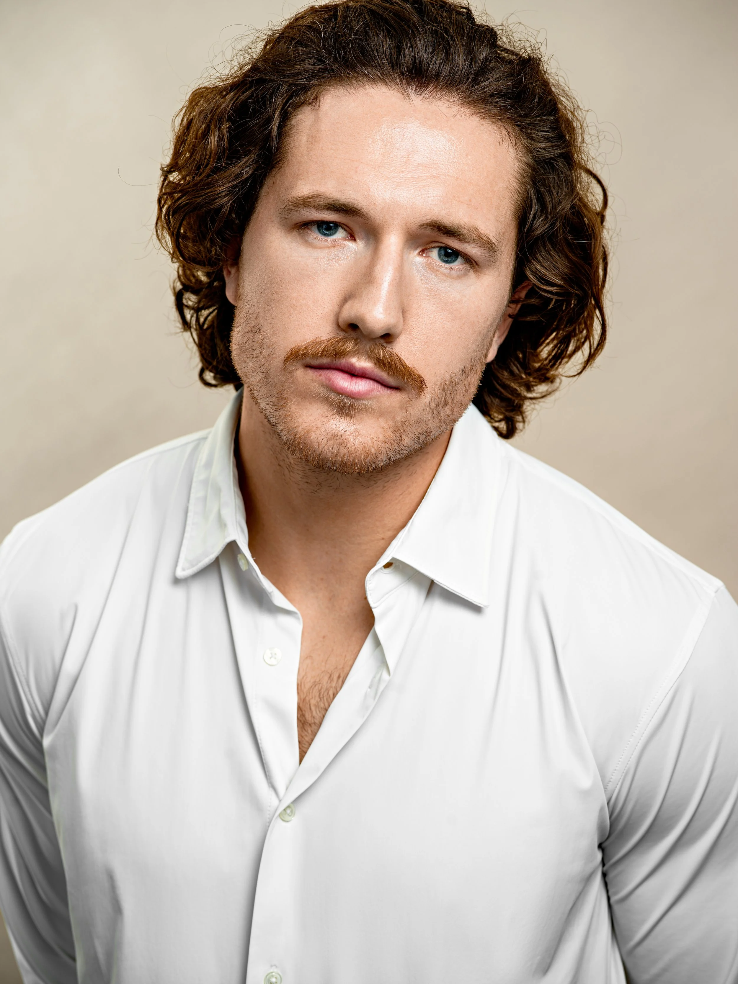 Close-up of a man with curly brown hair, blue eyes, and a light beard, wearing a white button-up shirt, against a plain background.