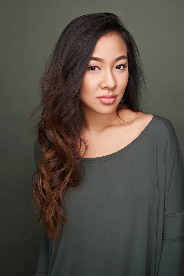 A young woman with long, wavy dark hair, light makeup, and a neutral expression, wearing a dark green long-sleeve top against a plain olive background.