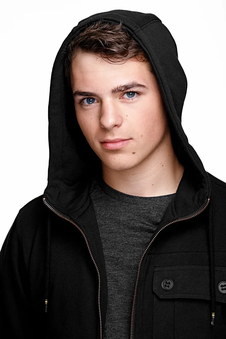 A young man with blue eyes and brown hair wearing a black hoodie with the hood up, layered over a dark gray shirt, against a white background.
