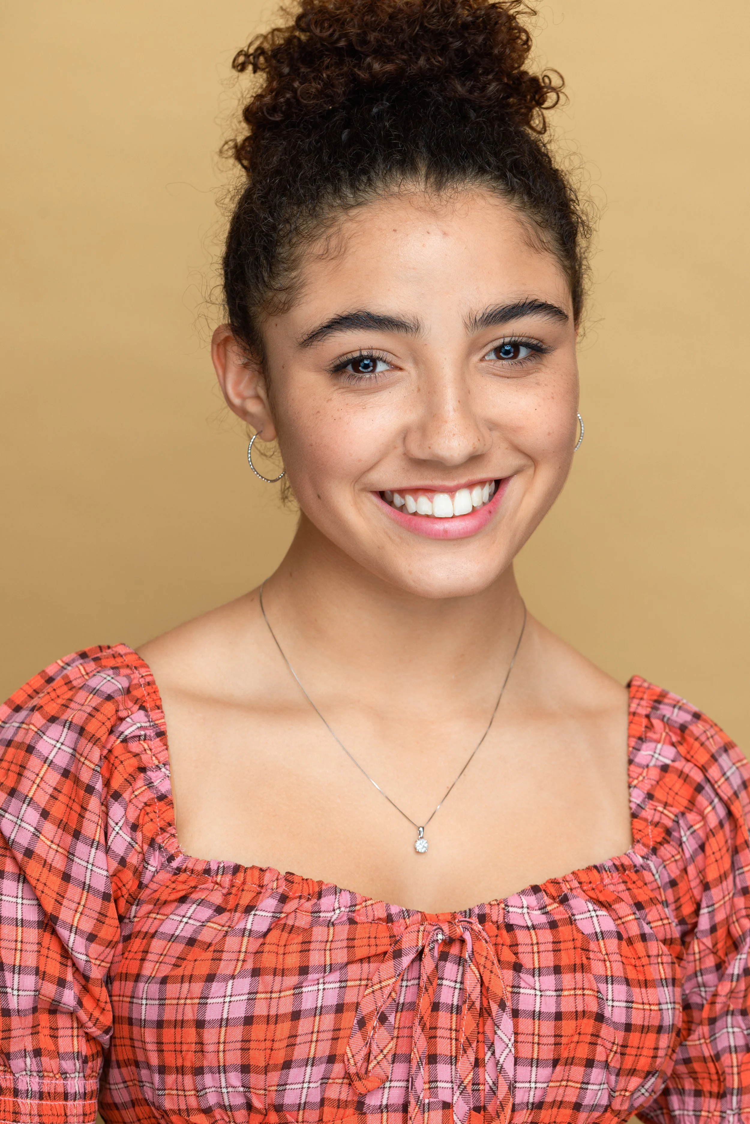 Young woman with curly hair styled in a top bun, wearing hoop earrings, a necklace with a pendant, and a red plaid blouse with a bow tie at the neckline, smiling against a beige background.