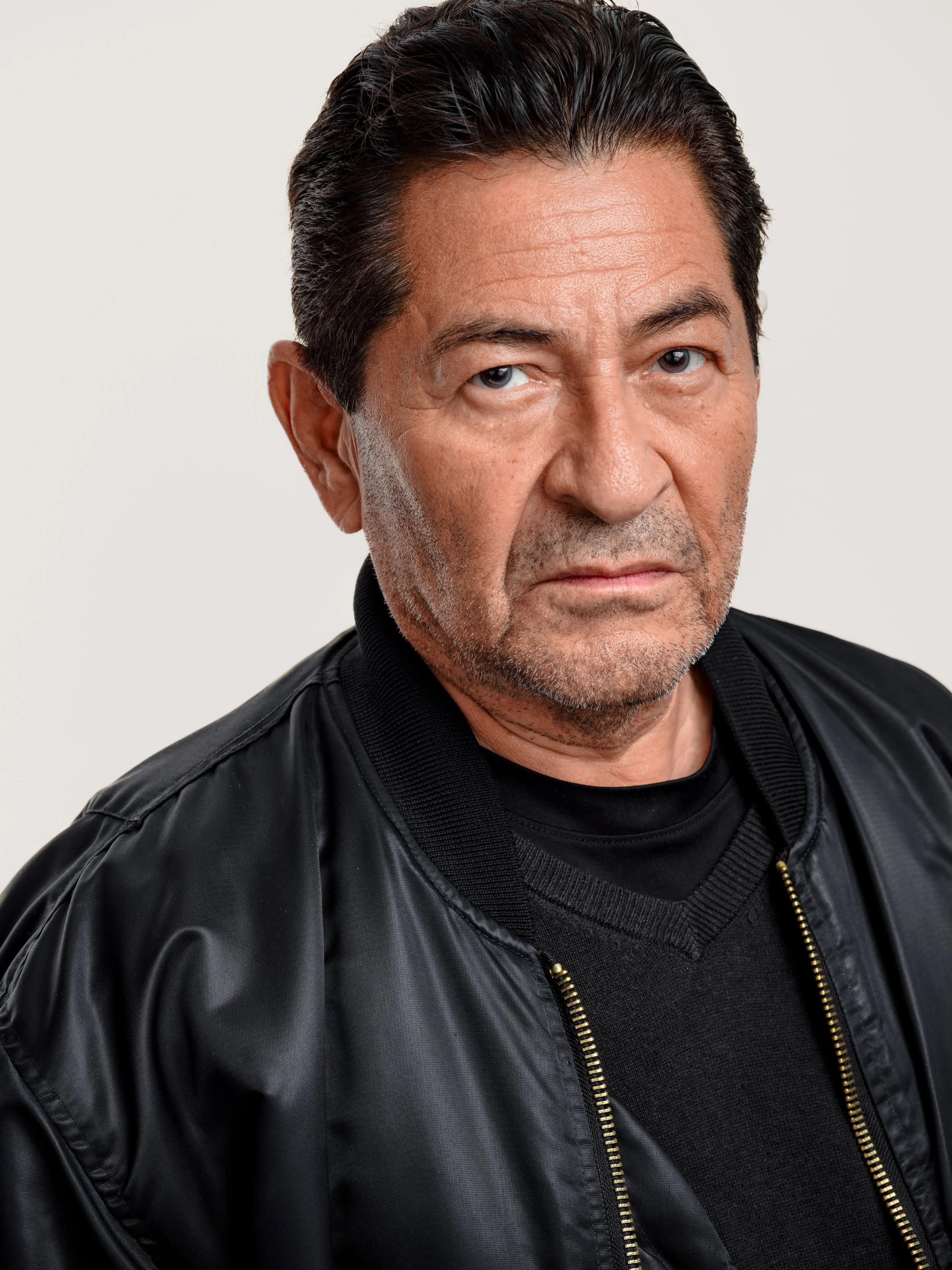 Close-up of a middle-aged man with dark hair and light skin, wearing a black leather jacket and black sweater, against a plain gray background.