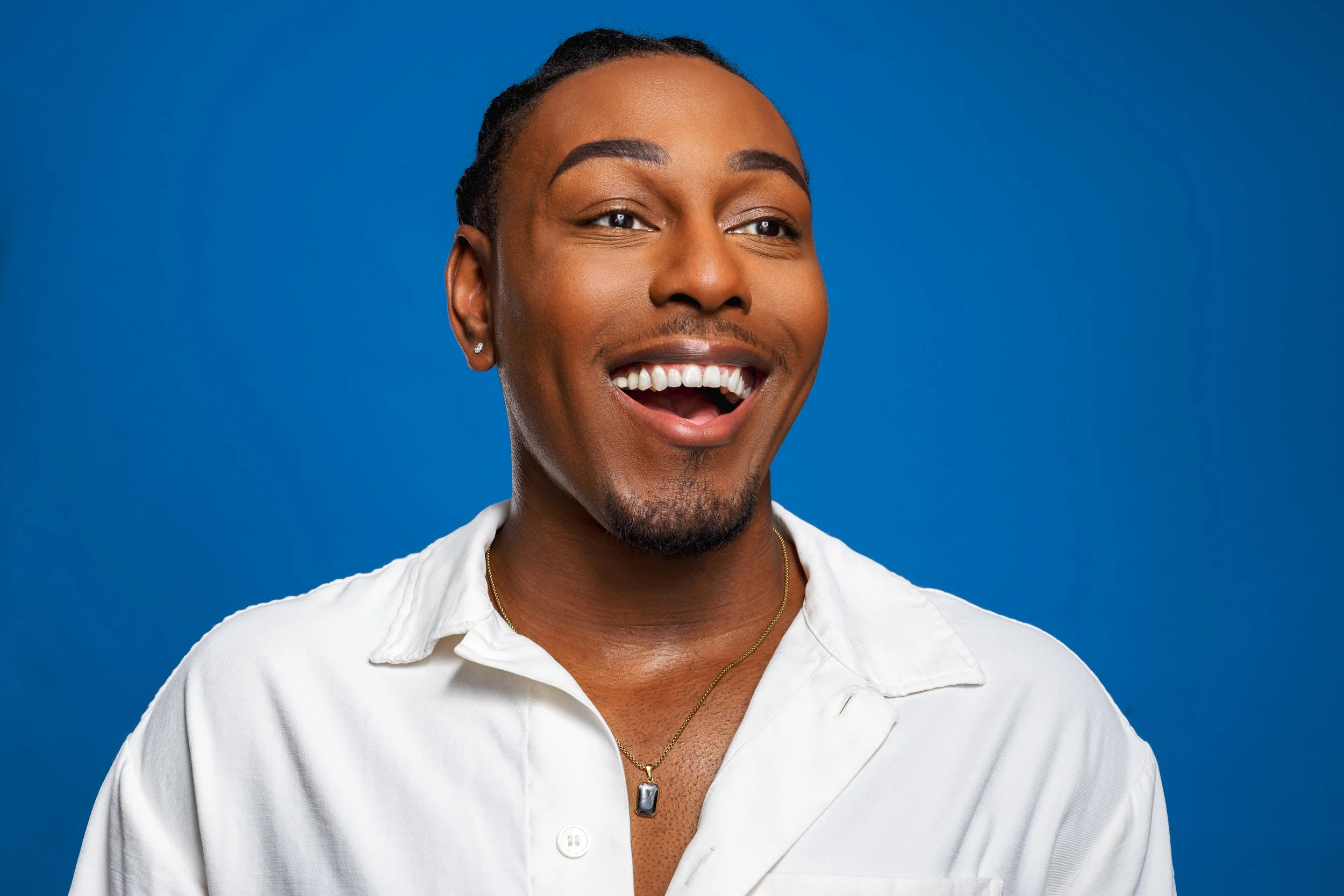 A smiling man with dark skin, short dreadlocked hair, wearing a white shirt, a pendant necklace, and small earrings, standing against a solid blue background.
