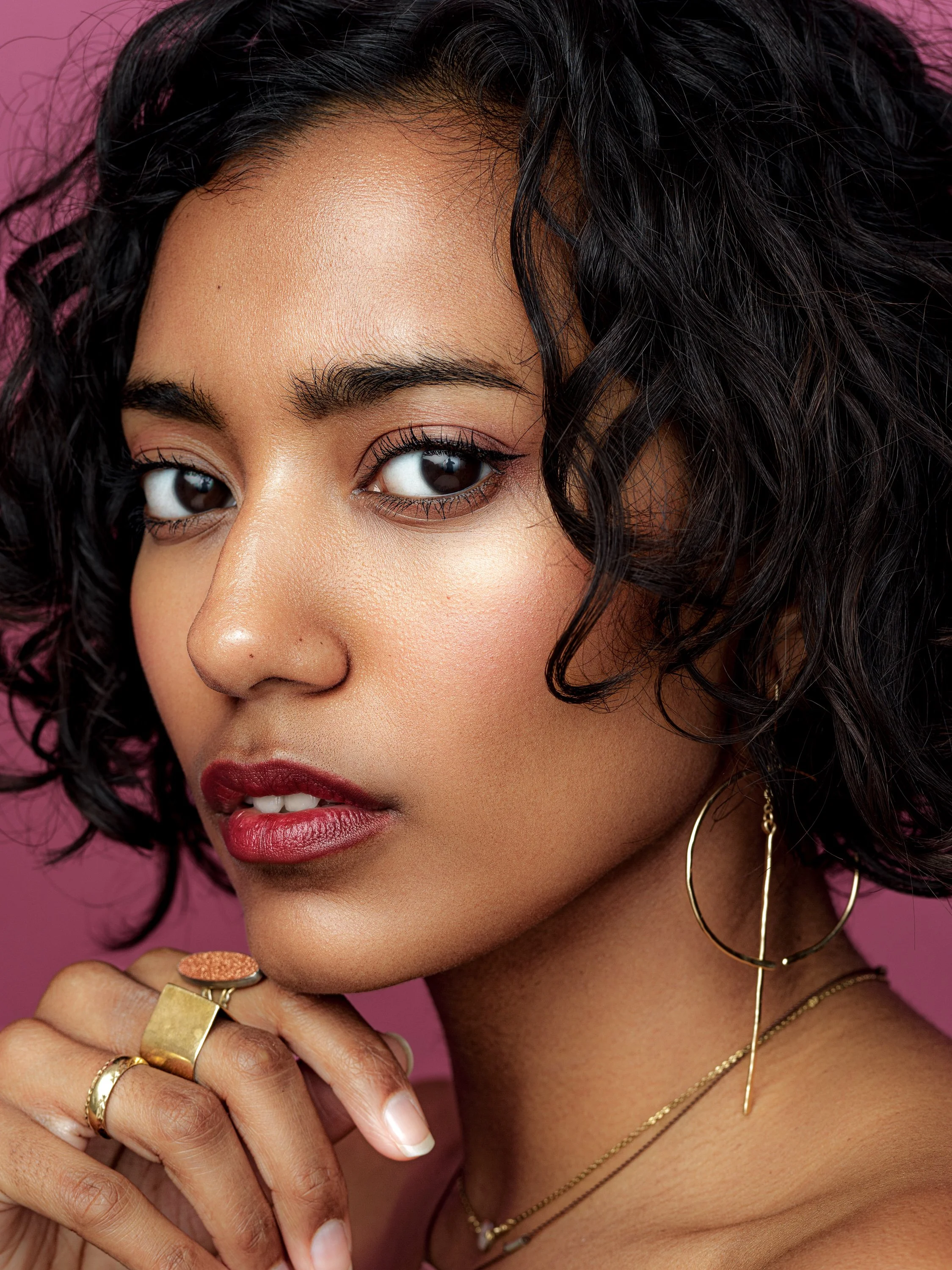 Close-up of a woman with dark, curly hair, wearing gold jewelry, bold makeup, and posing against a pink background.