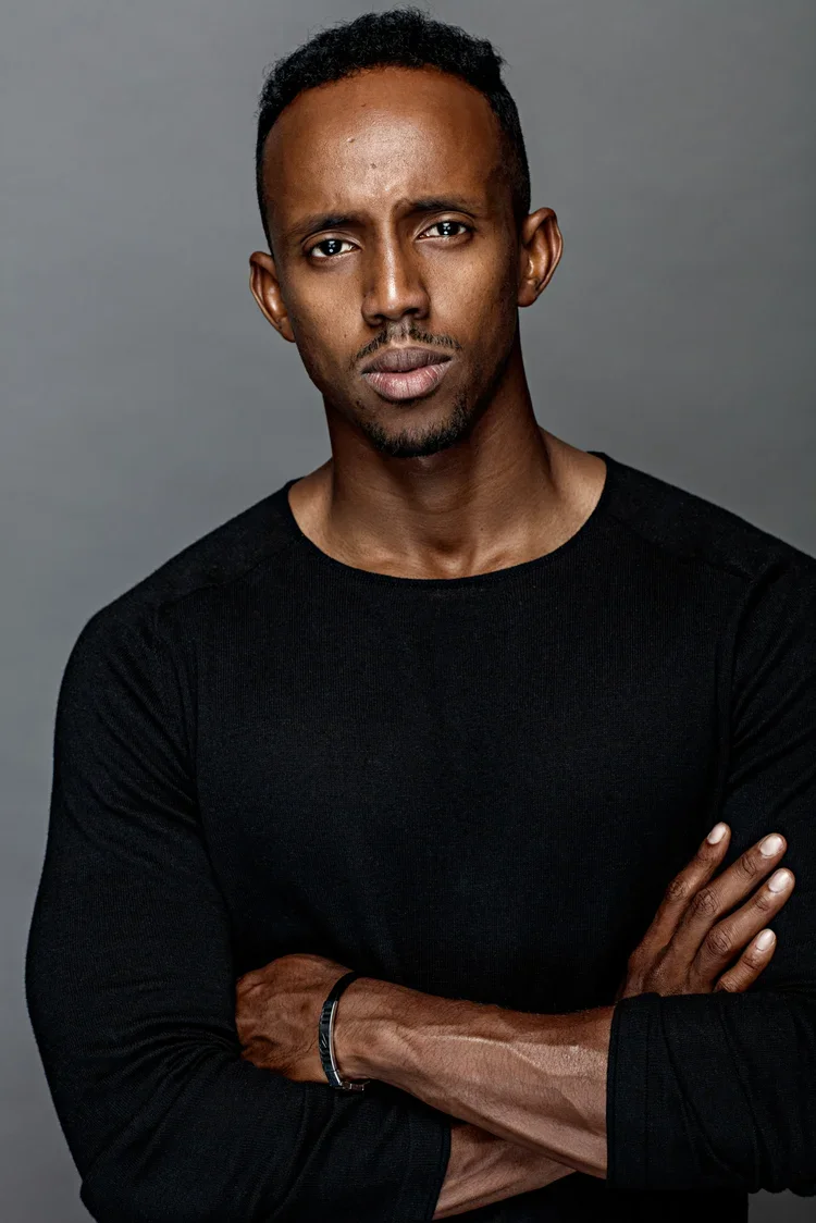 Portrait of a man with short, curly black hair, dark skin, wearing a black long-sleeve shirt, standing with arms crossed against a gray background.