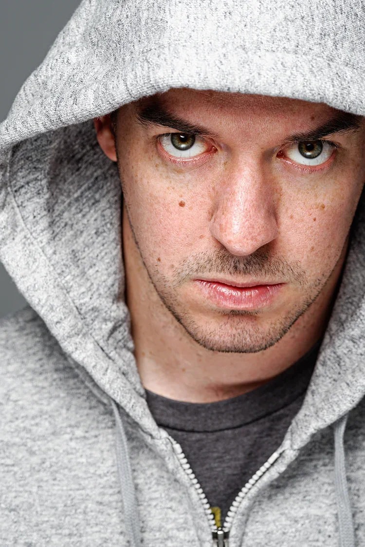 A close-up of a young man with blue eyes, freckles, and dark facial hair, wearing a gray hoodie with the hood over his head.