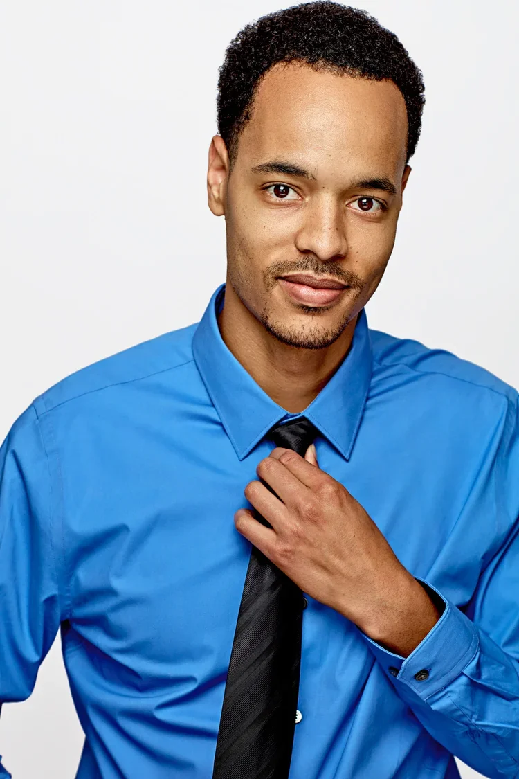 A young man with short curly hair and a goatee, wearing a blue dress shirt and a black tie, adjusting his tie with a confident smile against a plain white background.