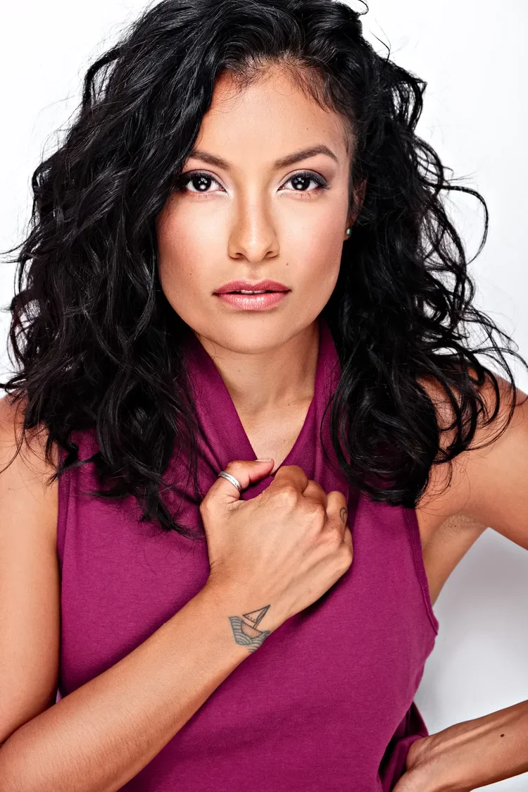 Portrait of a woman with curly black hair wearing a sleeveless purple top, holding her shirt collar with one hand, showing a tattoo of a sailboat on her wrist, against a plain white background.