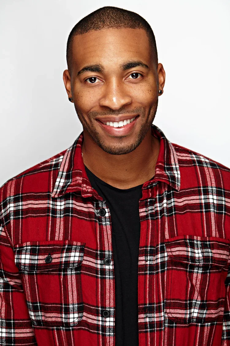 A young man with dark skin and short hair, smiling, wearing a red and black plaid shirt over a black T-shirt, against a white background.