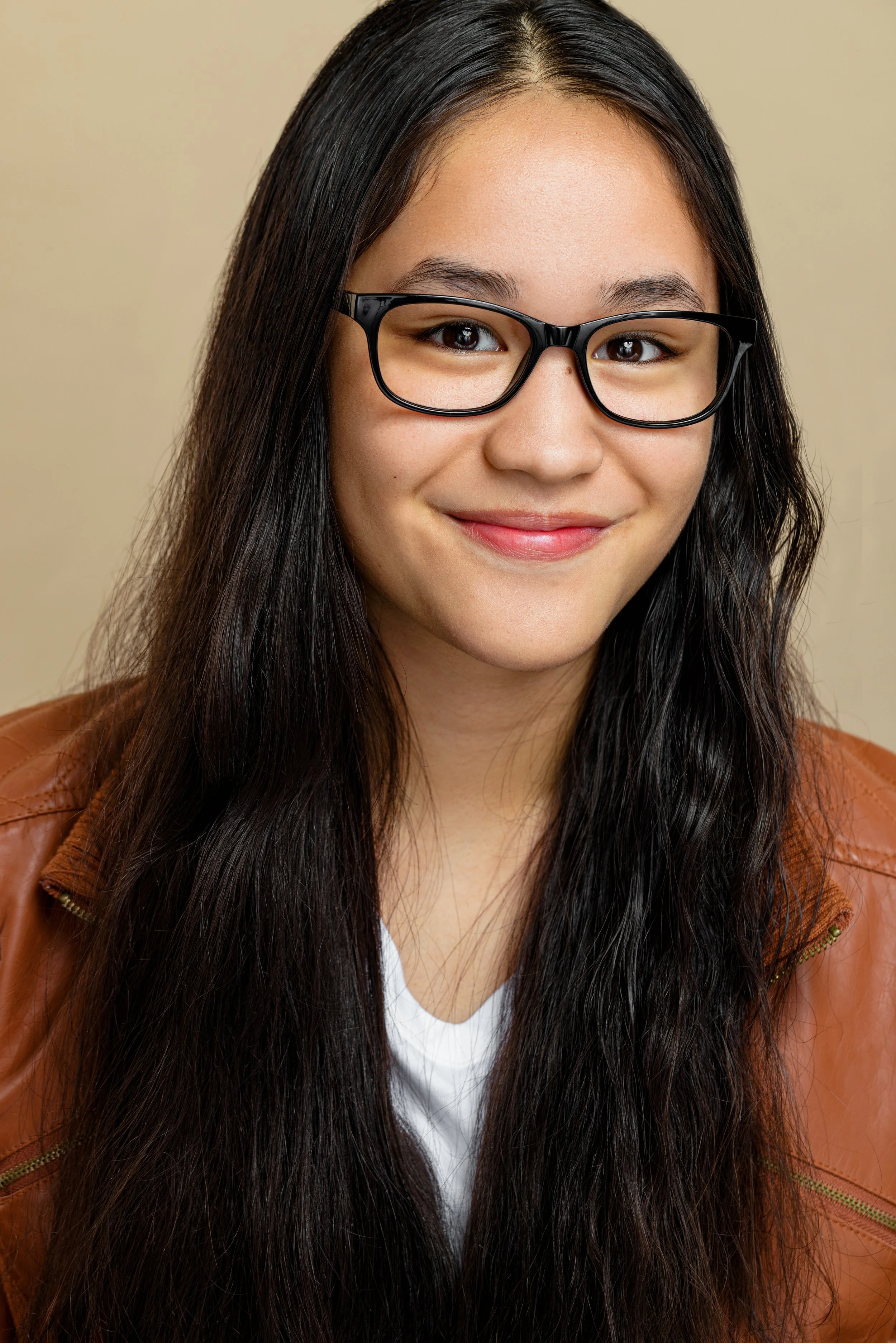 Close-up of a young woman with long dark hair wearing glasses and a brown leather jacket, smiling softly against a beige background.