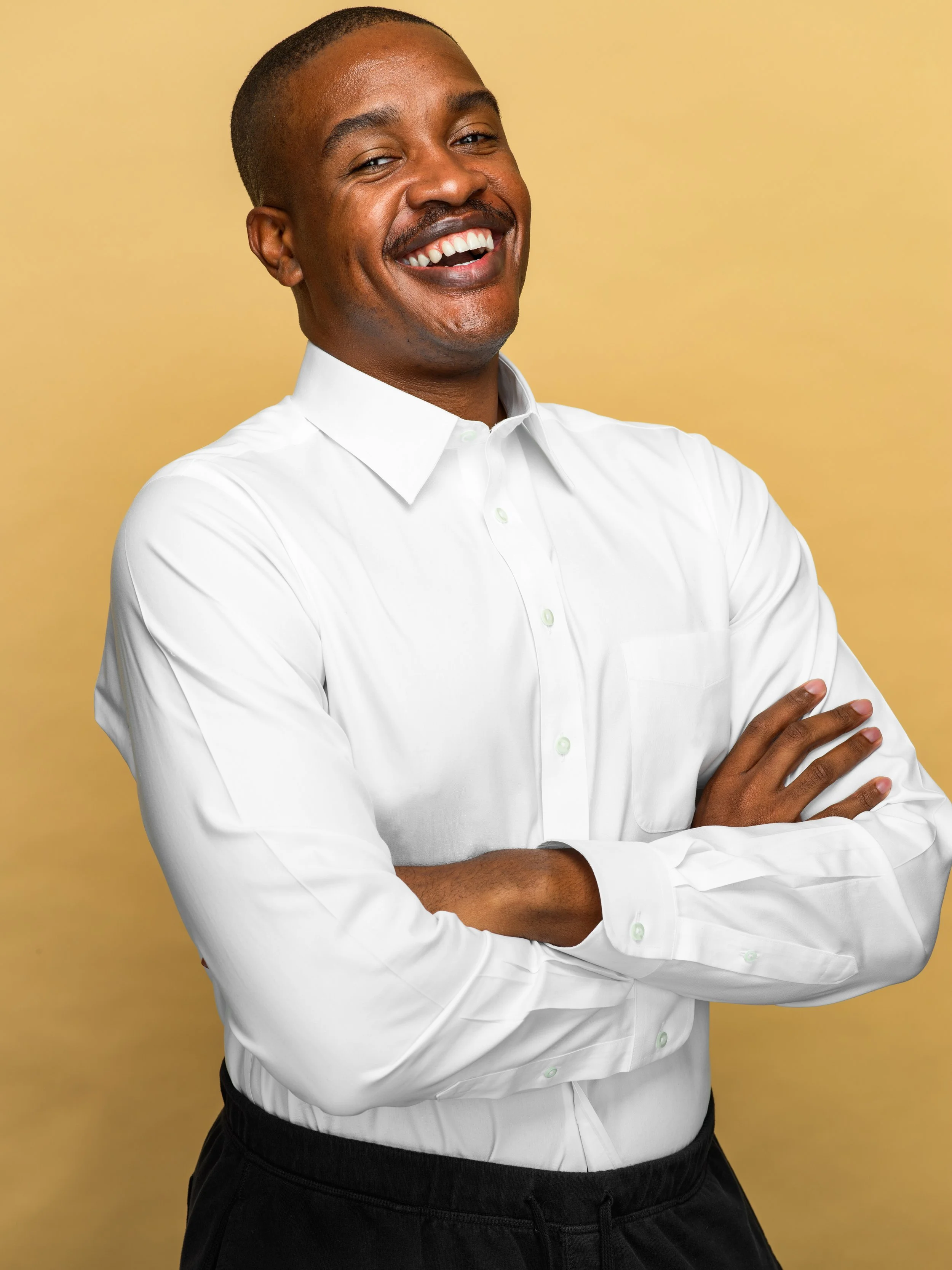 A smiling man with a mustache in a white dress shirt and black pants standing with arms crossed against a yellow background.