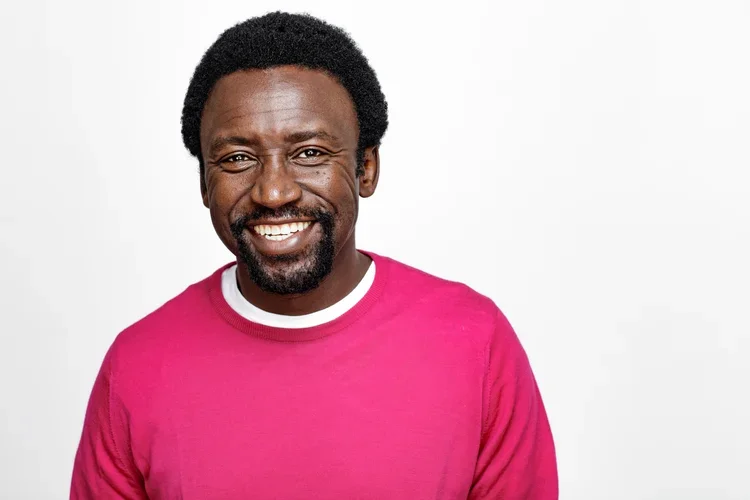 A smiling man with dark skin, black curly hair, and a beard, wearing a pink sweater against a plain white background.