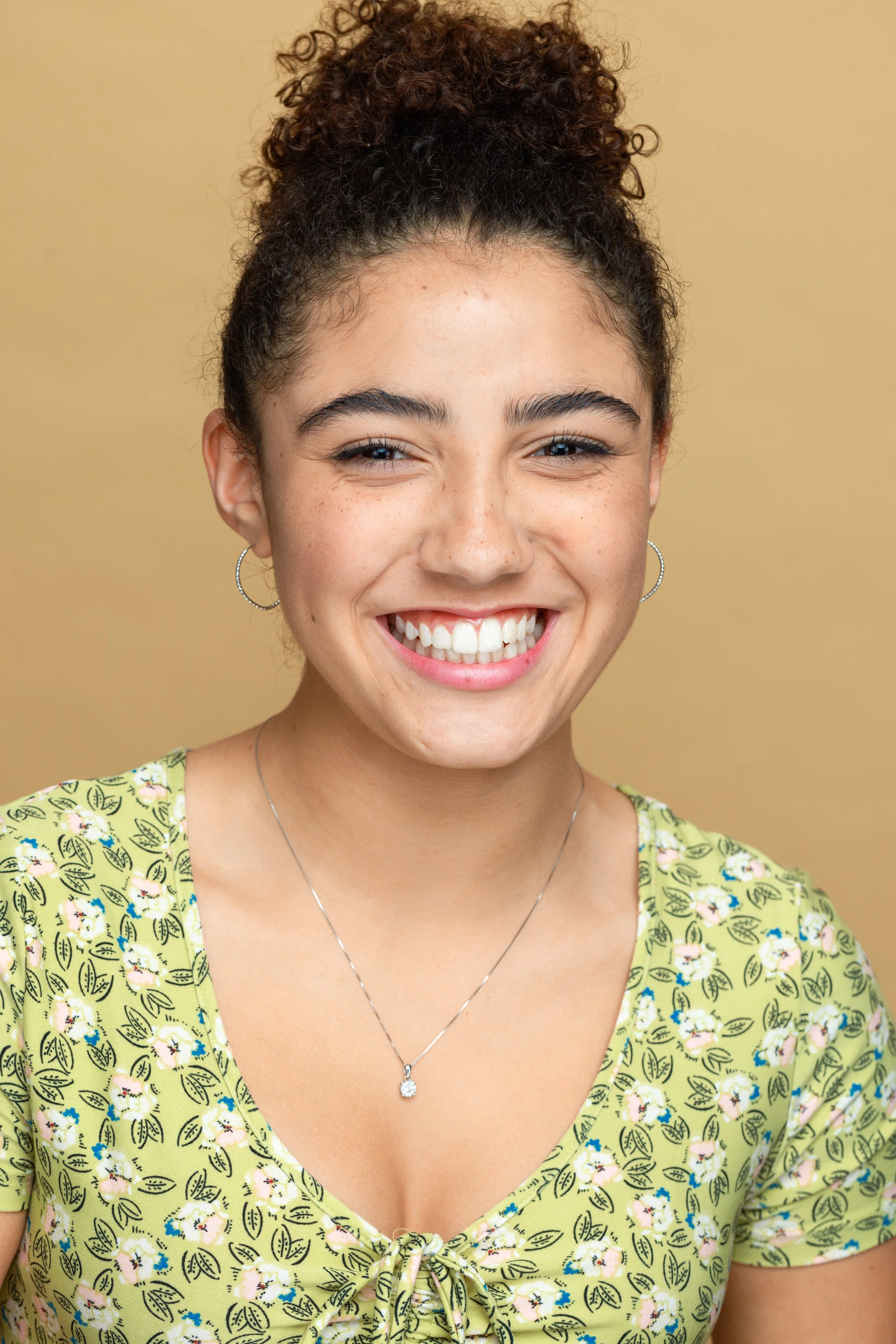 A young woman with curly brown hair styled in a high bun, smiling broadly and wearing a yellow dress with a floral pattern and a silver necklace with a pendant, standing against a beige background.