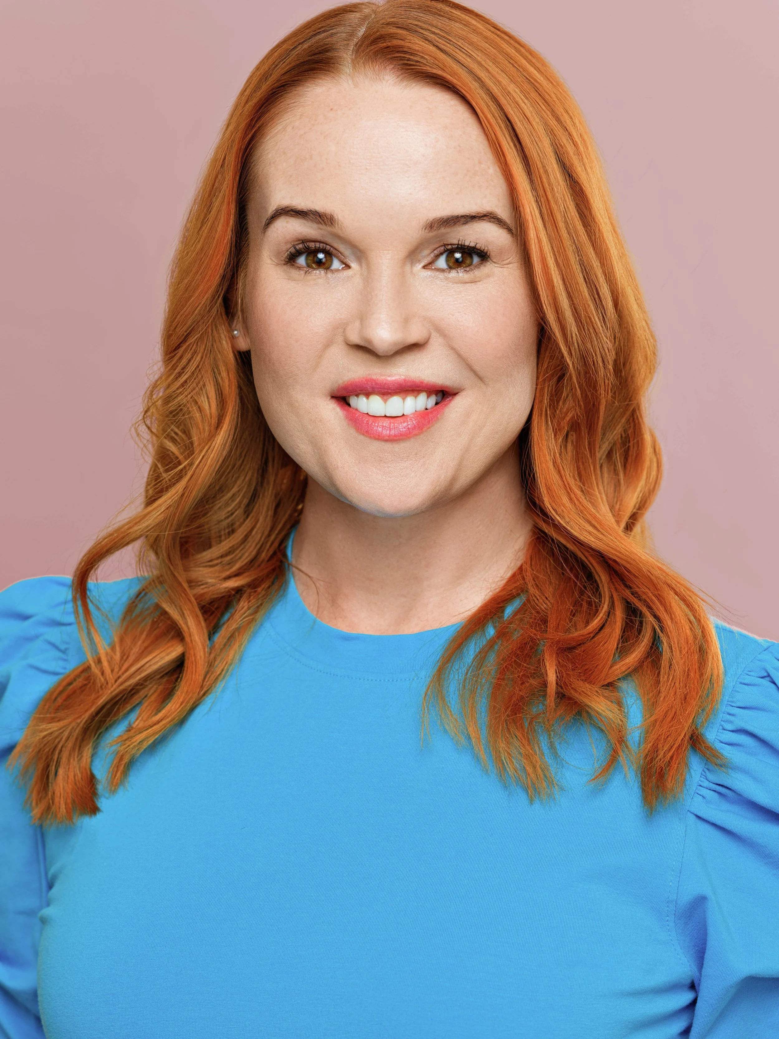 A smiling woman with red hair wearing a blue top against a pink background.