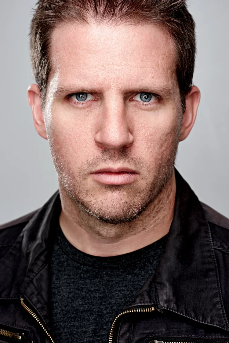 A close-up portrait of a man with short, dark hair, light skin, and piercing blue eyes, wearing a black jacket with a zipper.