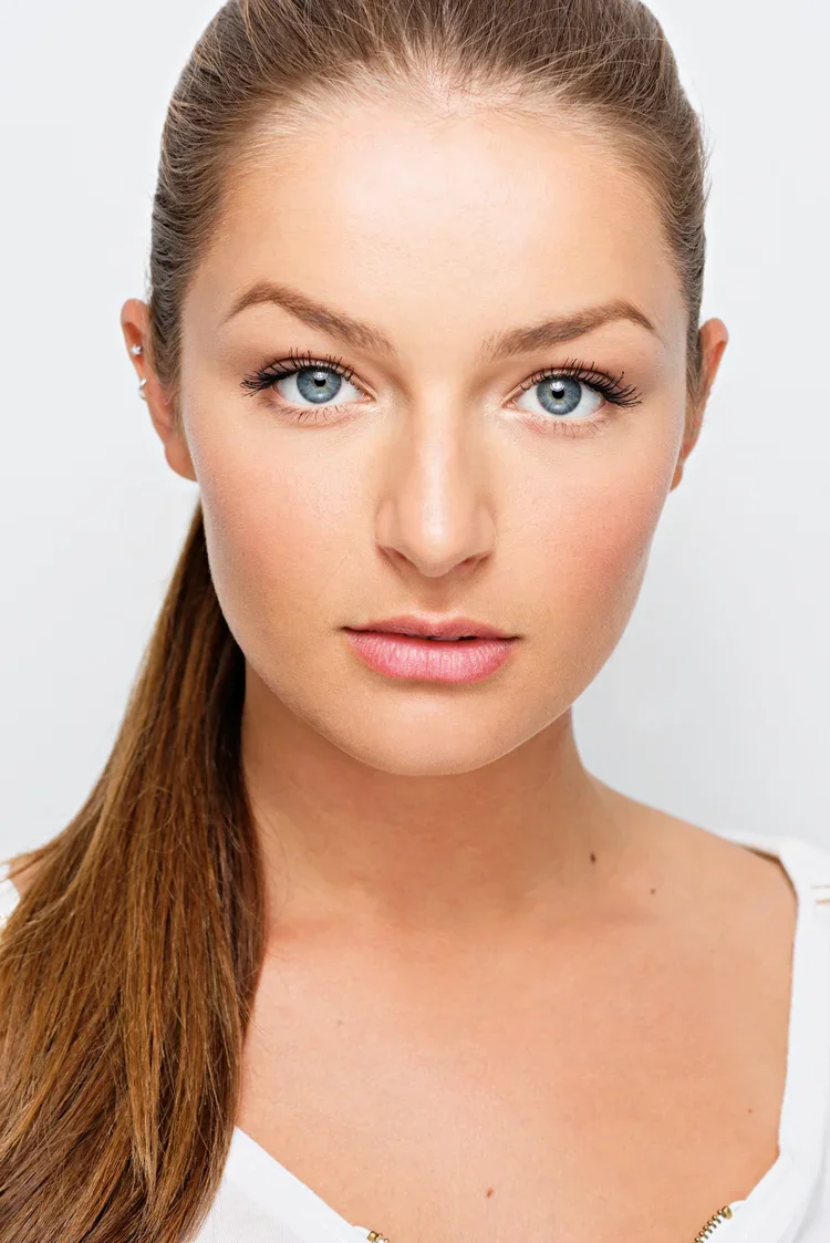 Close-up portrait of a young woman with blue eyes, light skin, and long brown hair tied back, wearing a white top.