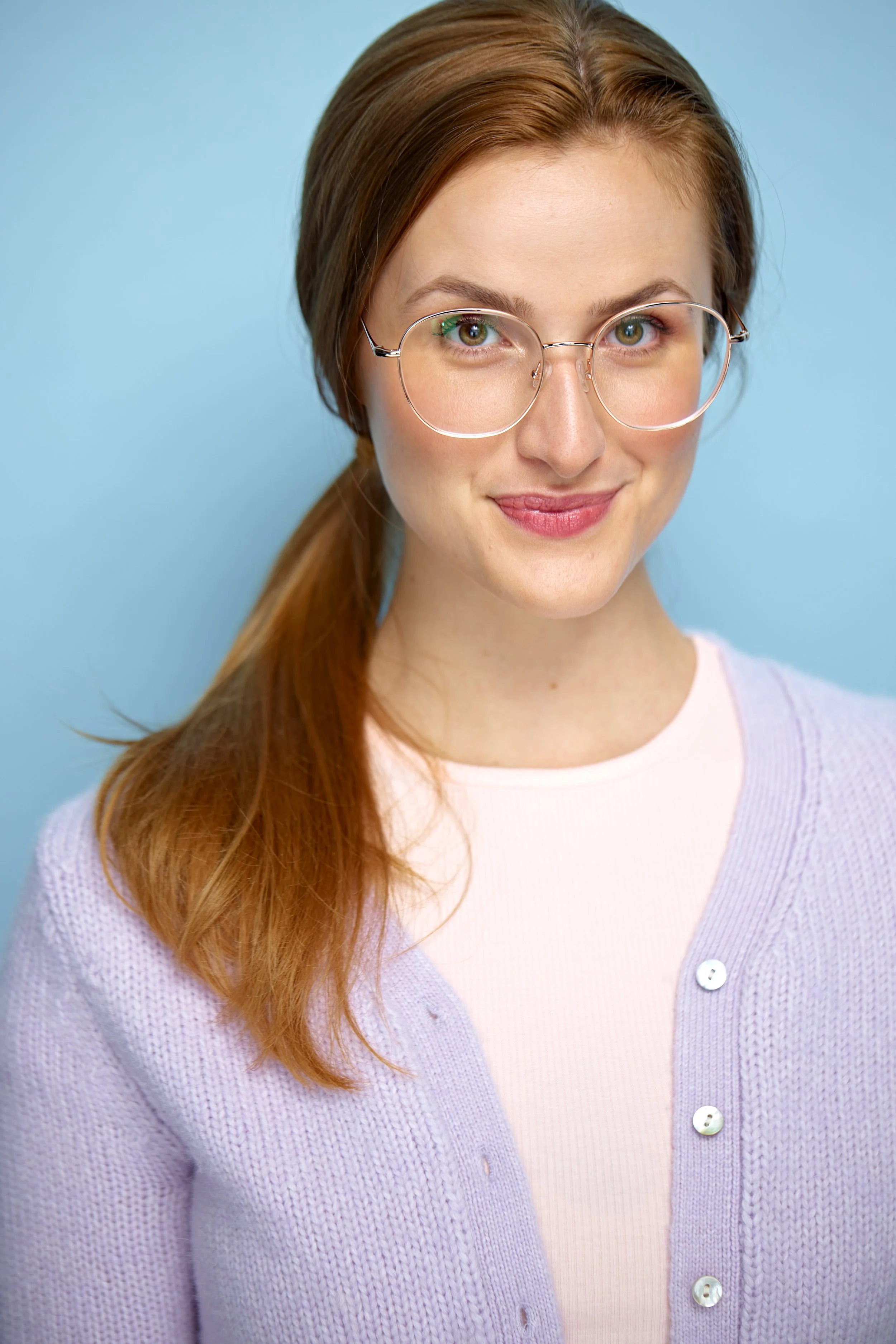 A young woman with light skin, red hair, and glasses smiling against a light blue background, wearing a lilac cardigan over a white top.