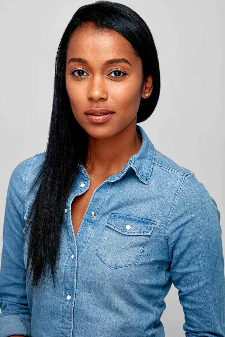 A young woman with straight black hair wearing a denim shirt against a plain grey background.