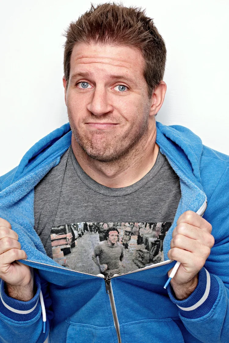 A man with light skin, blue eyes, and short brown hair wearing a gray T-shirt and a blue zip-up hoodie. He is lifting his hoodie to show a T-shirt with a picture of himself in a store aisle, looking down the aisle with a gray shirt. The man has a sli