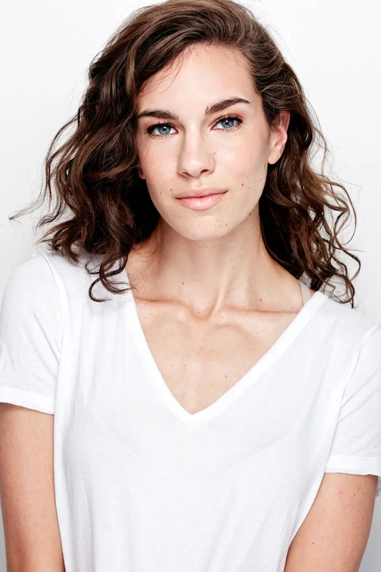 A young woman with light skin, blue eyes, and brown curly hair looking at the camera, wearing a white V-neck T-shirt, against a plain white background.