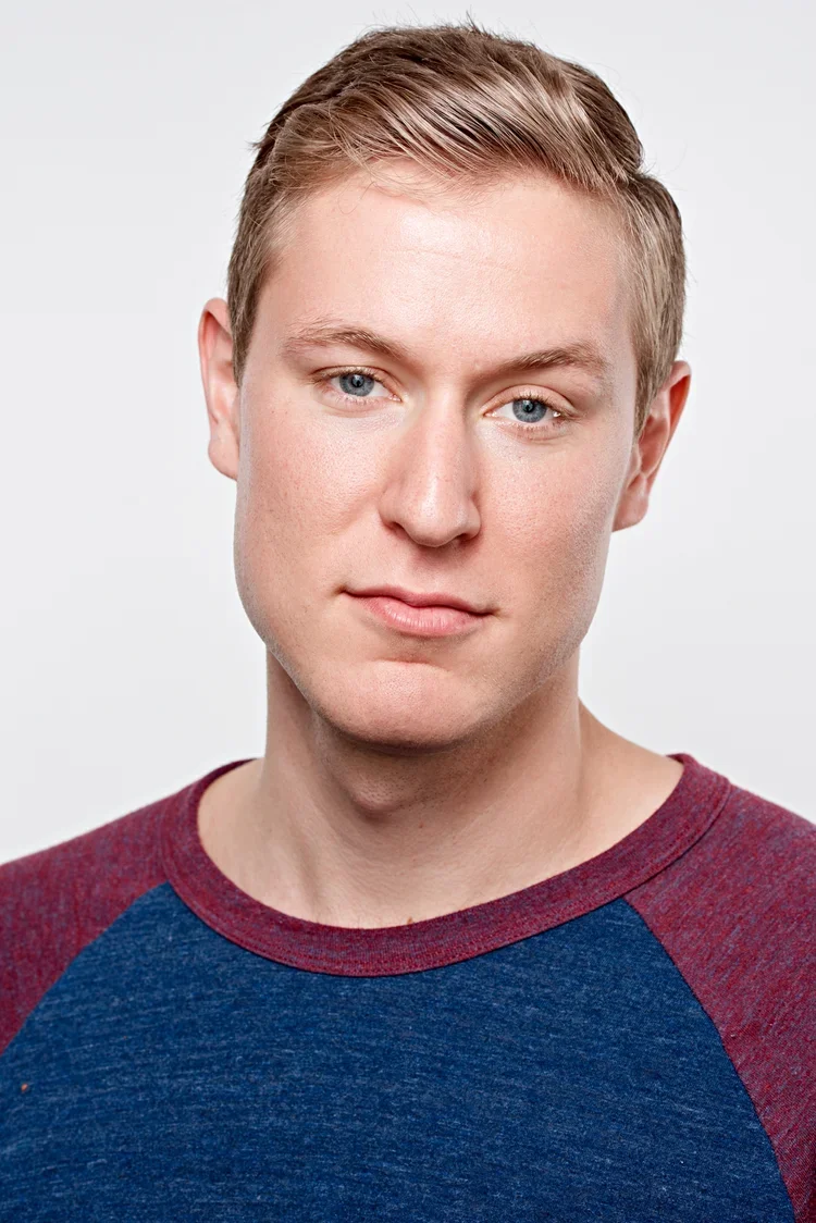 Portrait of a young man with short blond hair, blue eyes, and fair skin, wearing a maroon and blue casual shirt, against a plain white background.