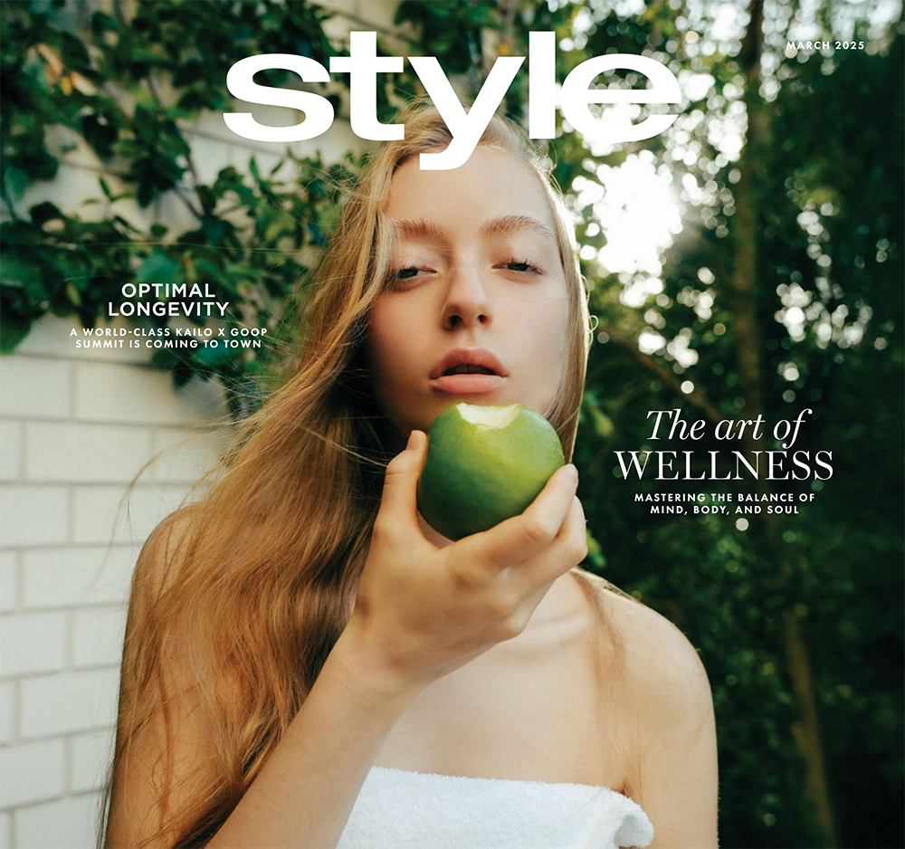 A woman with long blonde hair holding a green apple with a bite taken out of it, outdoors with greenery in the background.