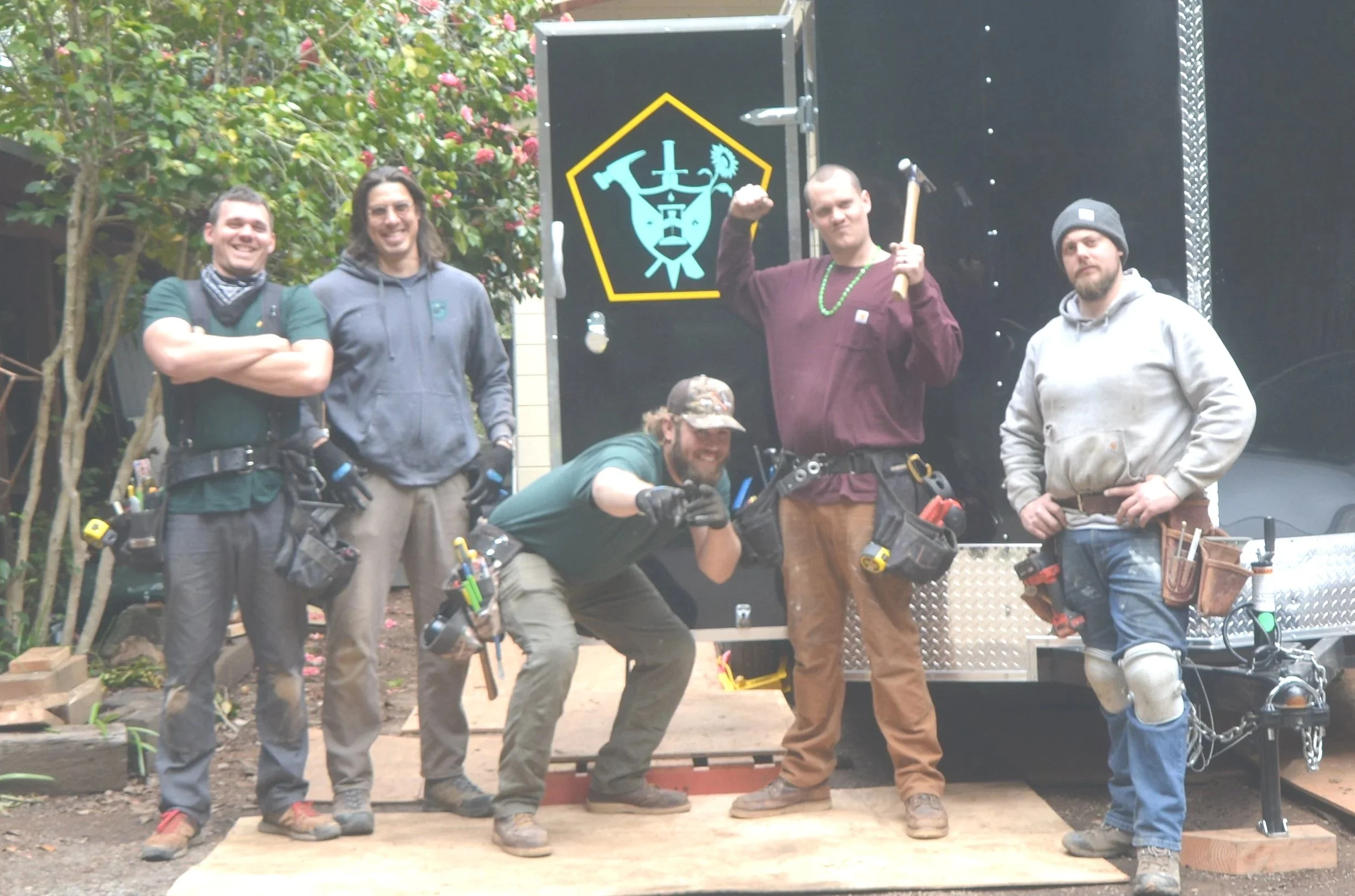 Group of five men with construction and maintenance tools posing outdoors in front of a black trailer with a logo. One man is flexing his arm, another crouching and pointing, and others stand with hands on hips or crossed arms.