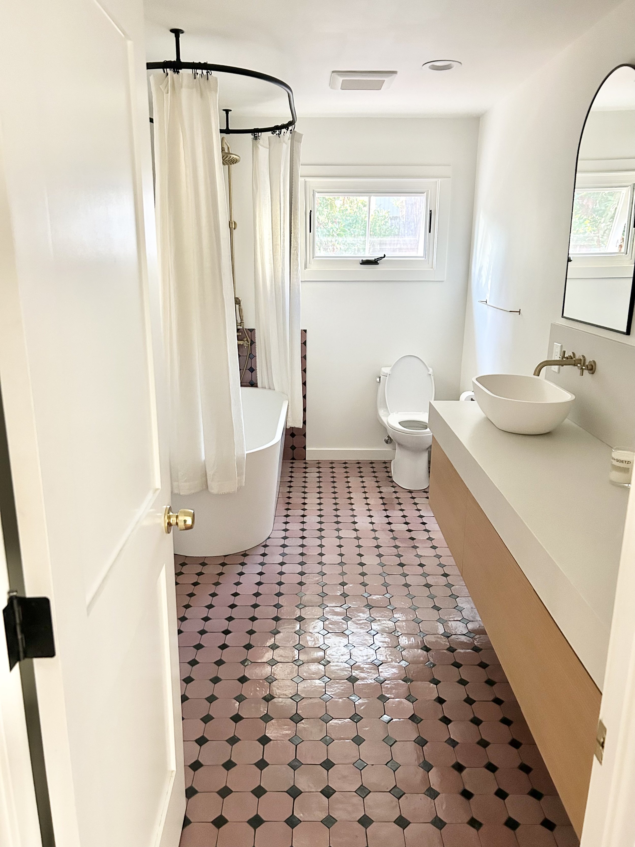 A clean, modern bathroom with pink and black hexagonal tile flooring, white walls, a window, a shower with a curtain, a white toilet, a floating vanity with a vessel sink, and a mirror.