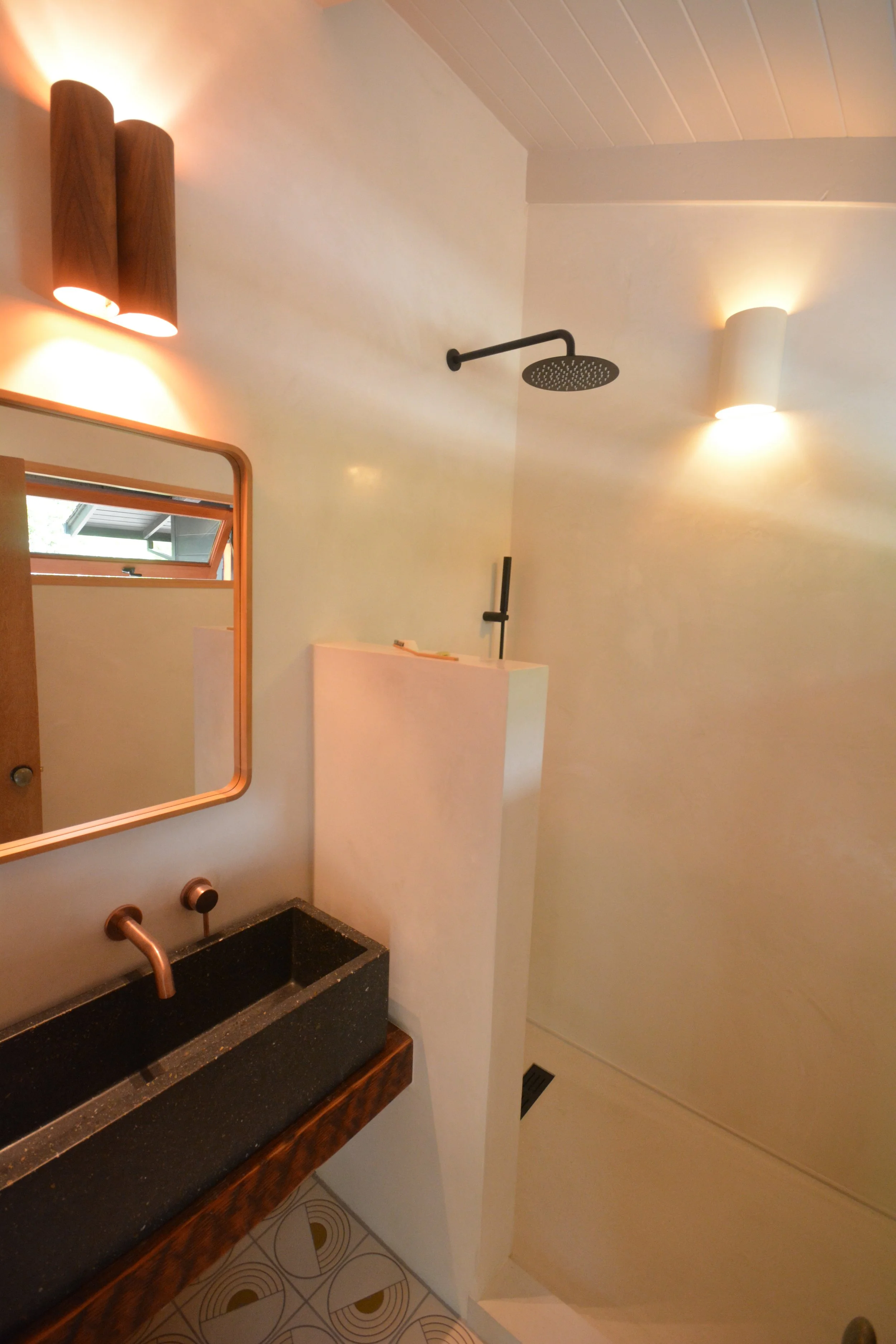 Modern bathroom with black stone sink, wooden mirror frame, and dual wall-mounted light fixtures. Shower area with black ceiling rain shower and a small window.
