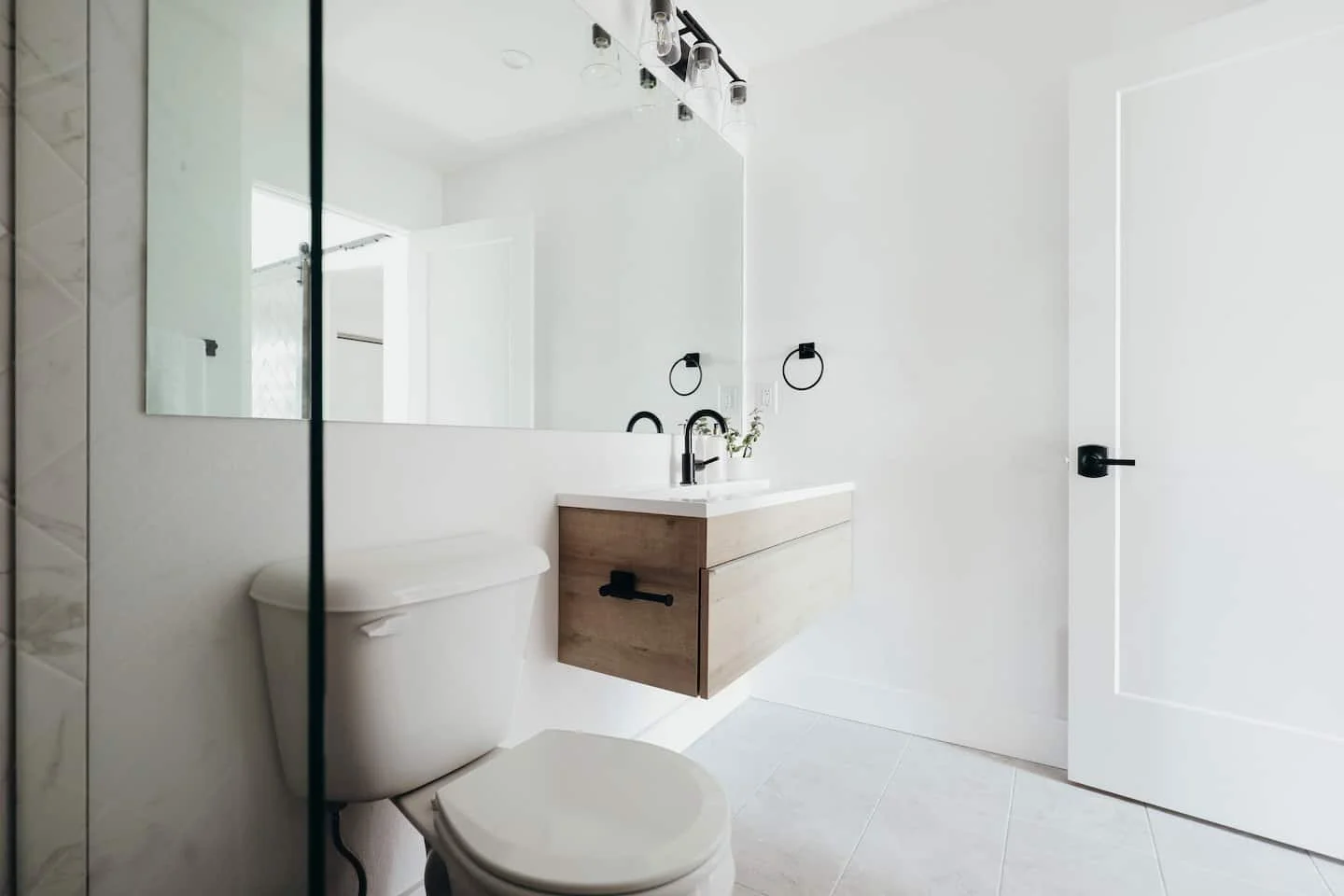 Minimalist white bathroom with floating wooden vanity, black fixtures, and a large mirror, featuring a toilet and a door.