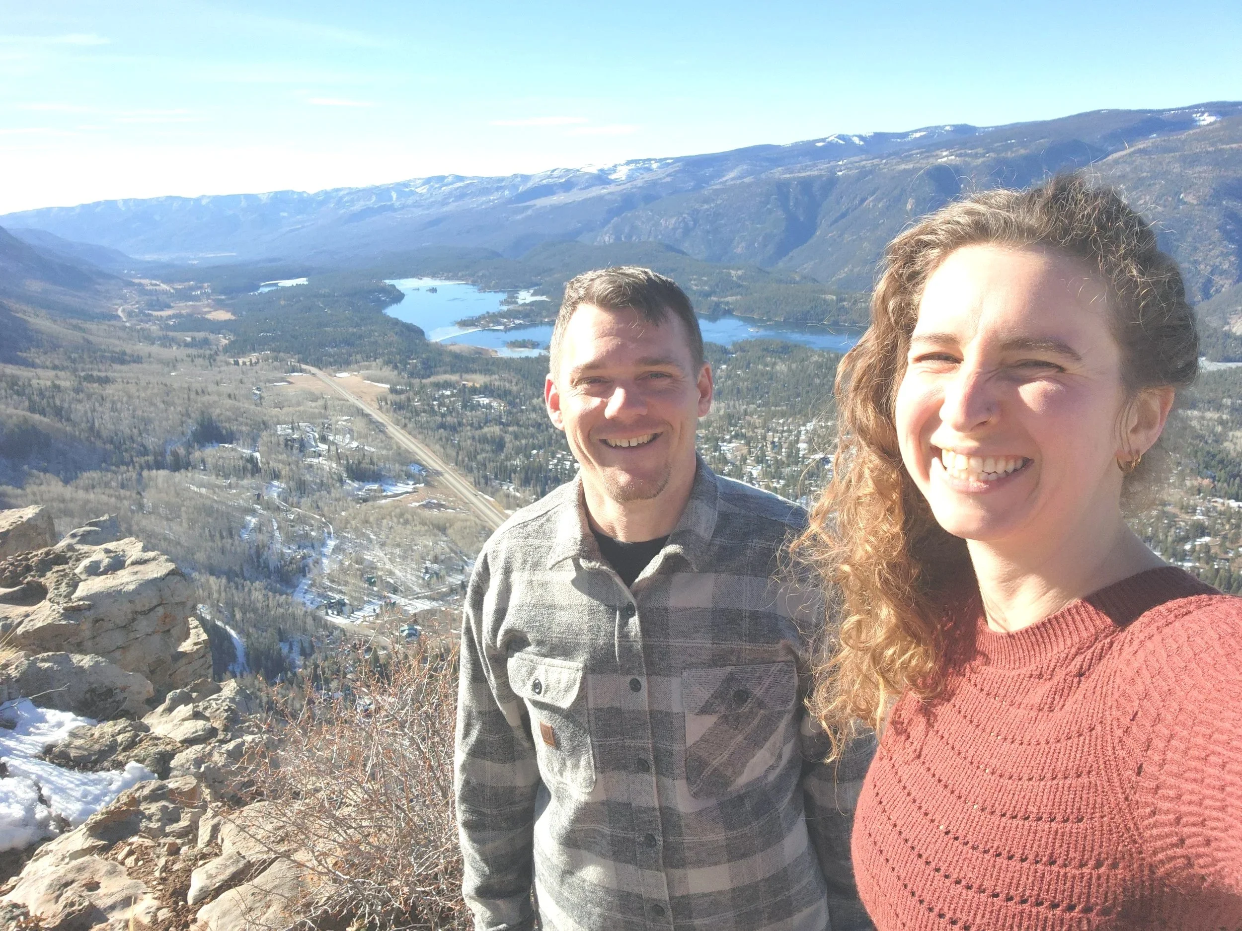 A smiling man and woman taking a selfie on a mountain overlook with a scenic view of lakes, mountains, and a small town below on a clear day.