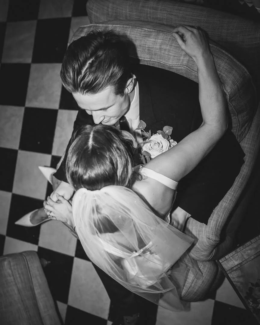 Black and white photo of a man in a suit and a woman in a wedding dress sharing a kiss while seated on a couch
