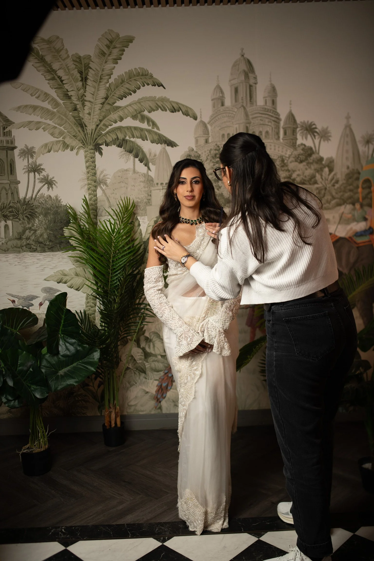 A woman in a white ornate dress and jewelry poses for a photo with another woman, who is adjusting her dress, in front of a tropical-themed mural with palm trees and a palace. Potted plants are on the floor, and there is black and white checkered tile at the bottom.