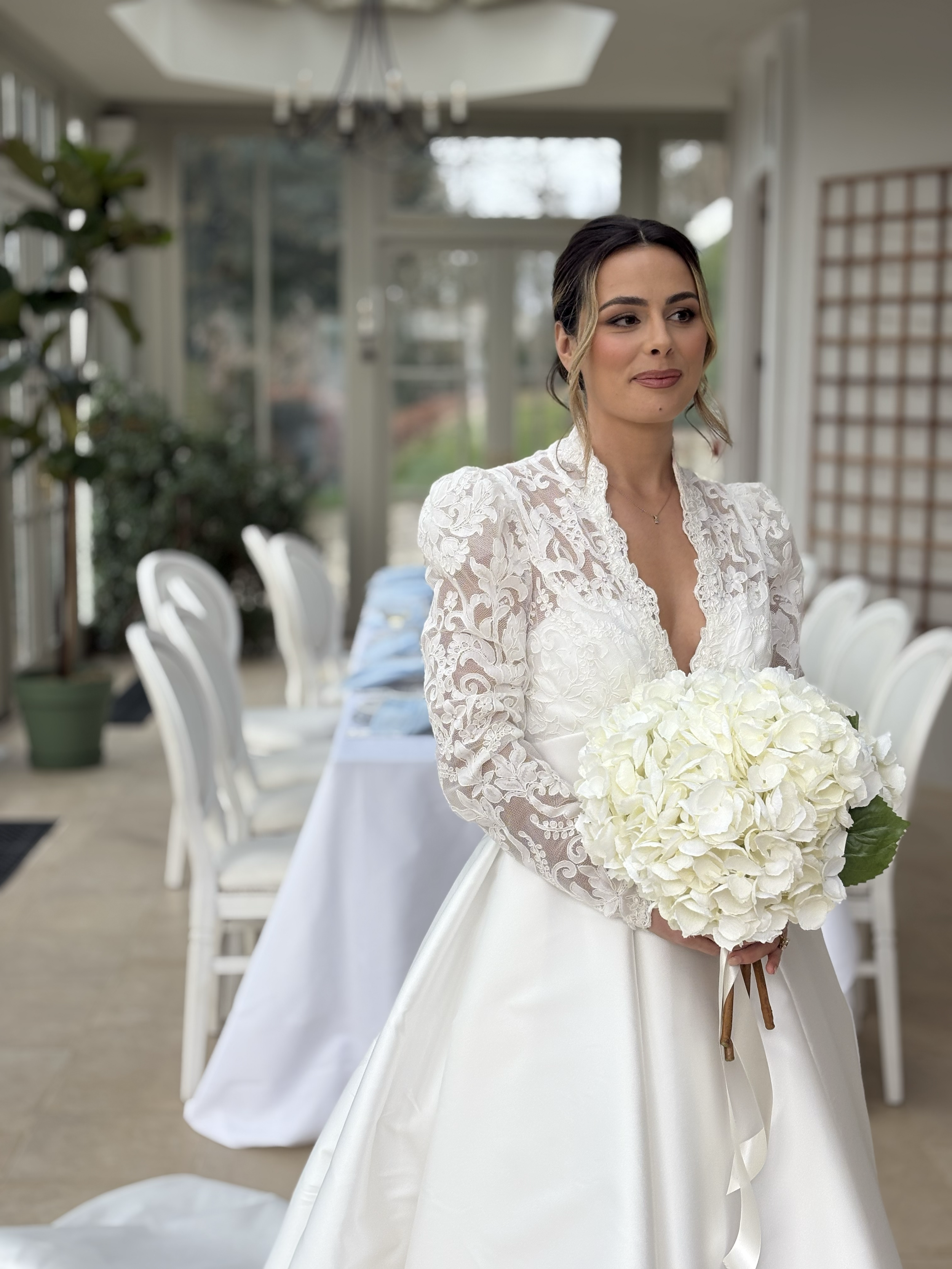 A bride in a white wedding dress holding a bouquet of white hydrangeas, standing in a bright sunroom with white chairs along a dining table.