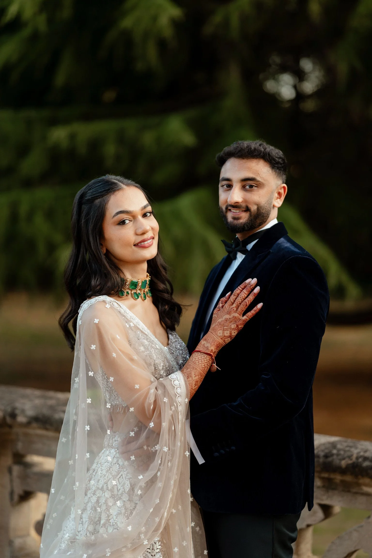 A couple dressed in wedding attire standing outdoors in front of green trees, smiling at the camera. The woman wears a white embroidered dress, a gold and green necklace, and has henna on her hands, while the man wears a black tuxedo with a bow tie.