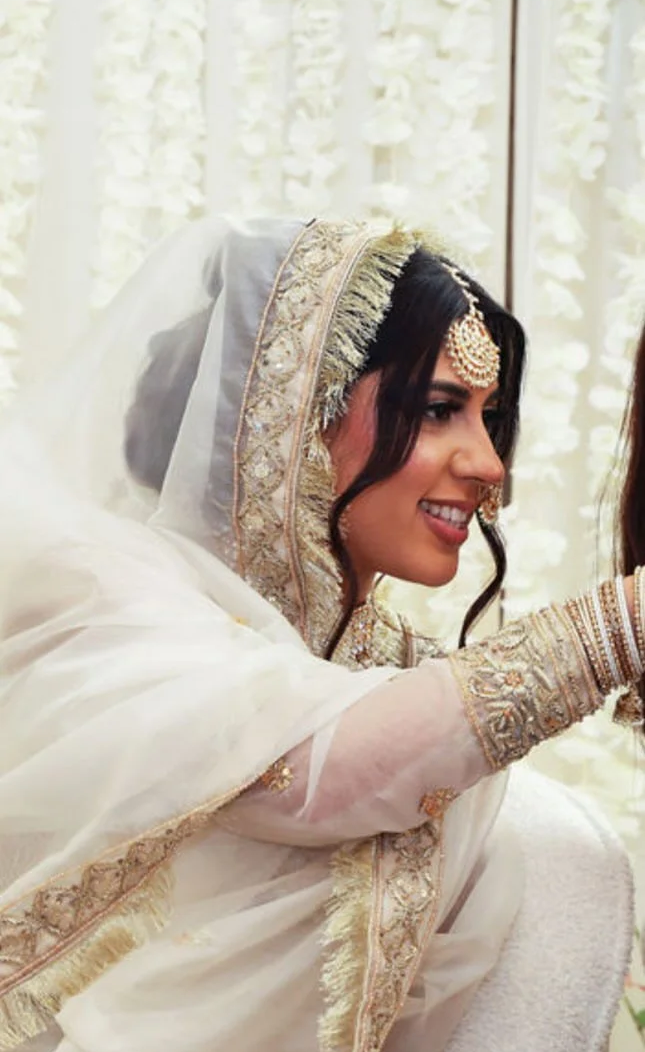A woman wearing traditional wedding attire, including a cream-colored dress with gold embroidery, jewelry, and a veil, smiling while sitting indoors with a floral backdrop.