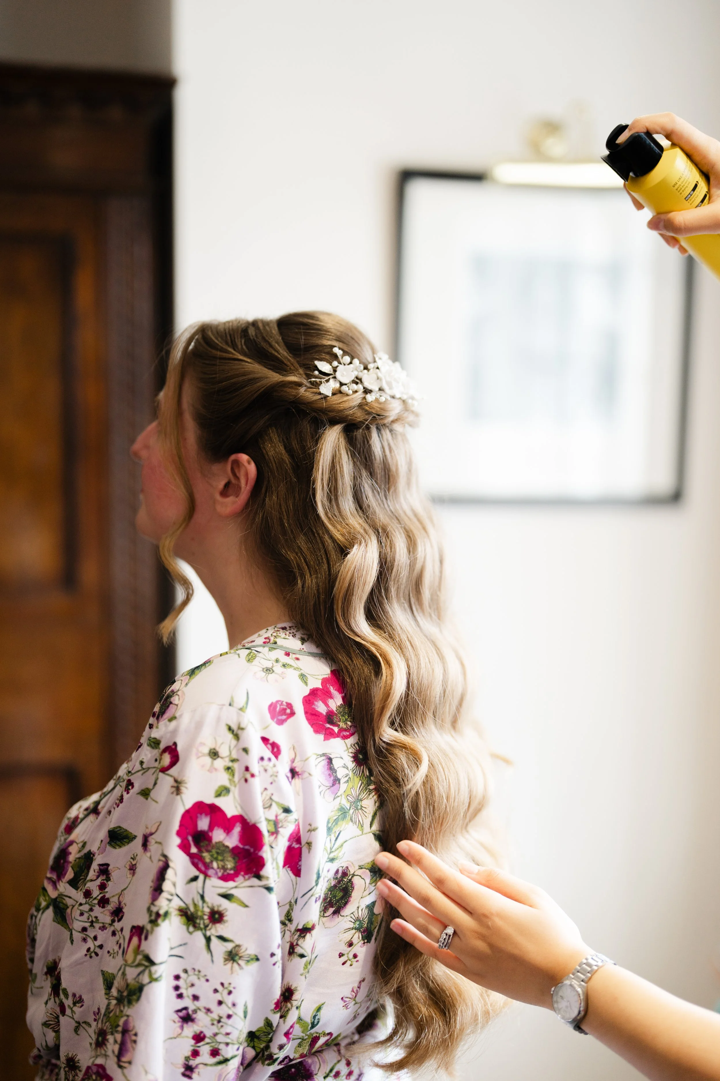 A woman with long blonde hair styled with a floral hairpiece is getting her hair styled, while someone else’s hand spritzes her hair with hair spray.