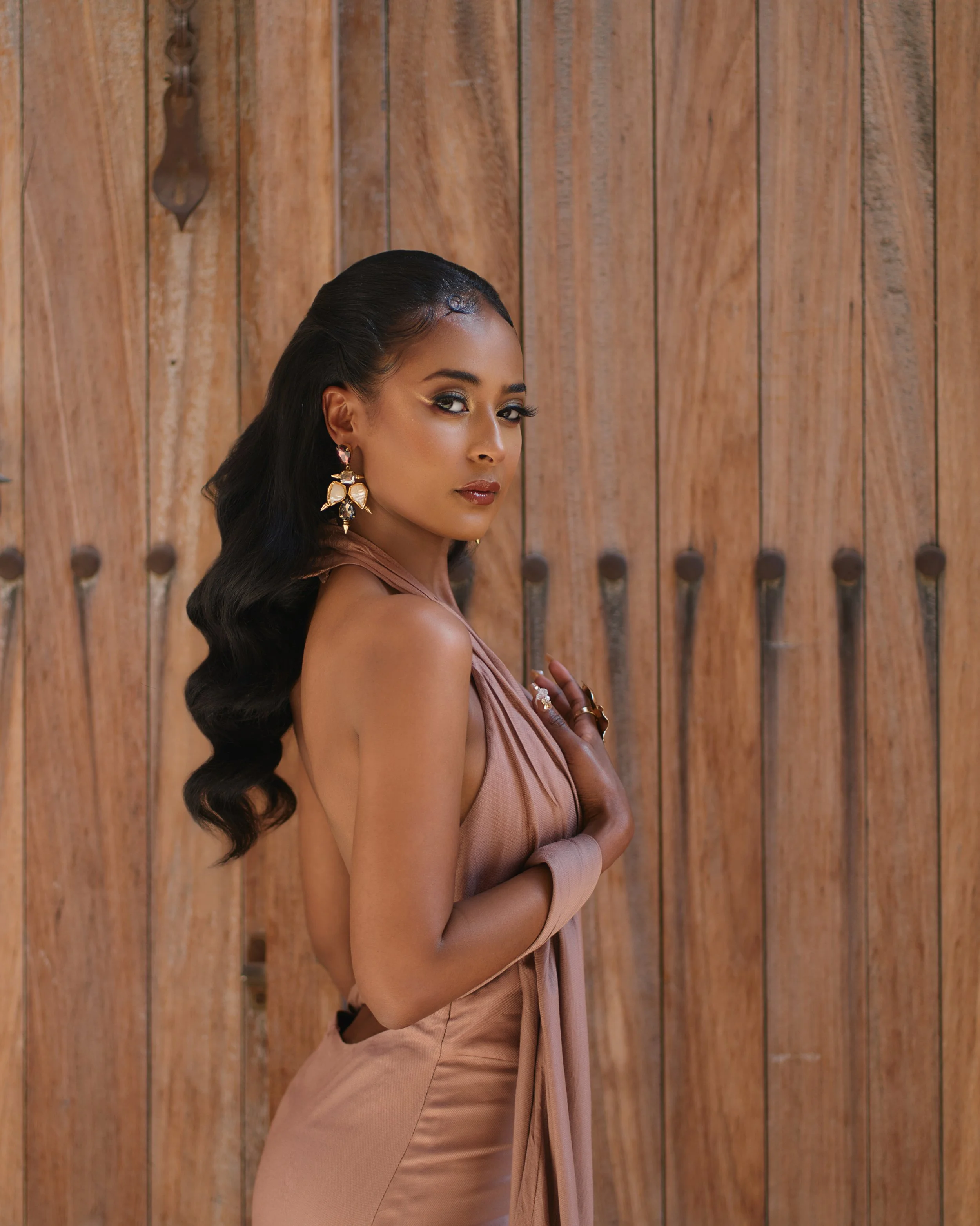 A woman with long black hair styled in waves, wearing ornate gold earrings, posed in front of a wooden fence with a confident expression.
