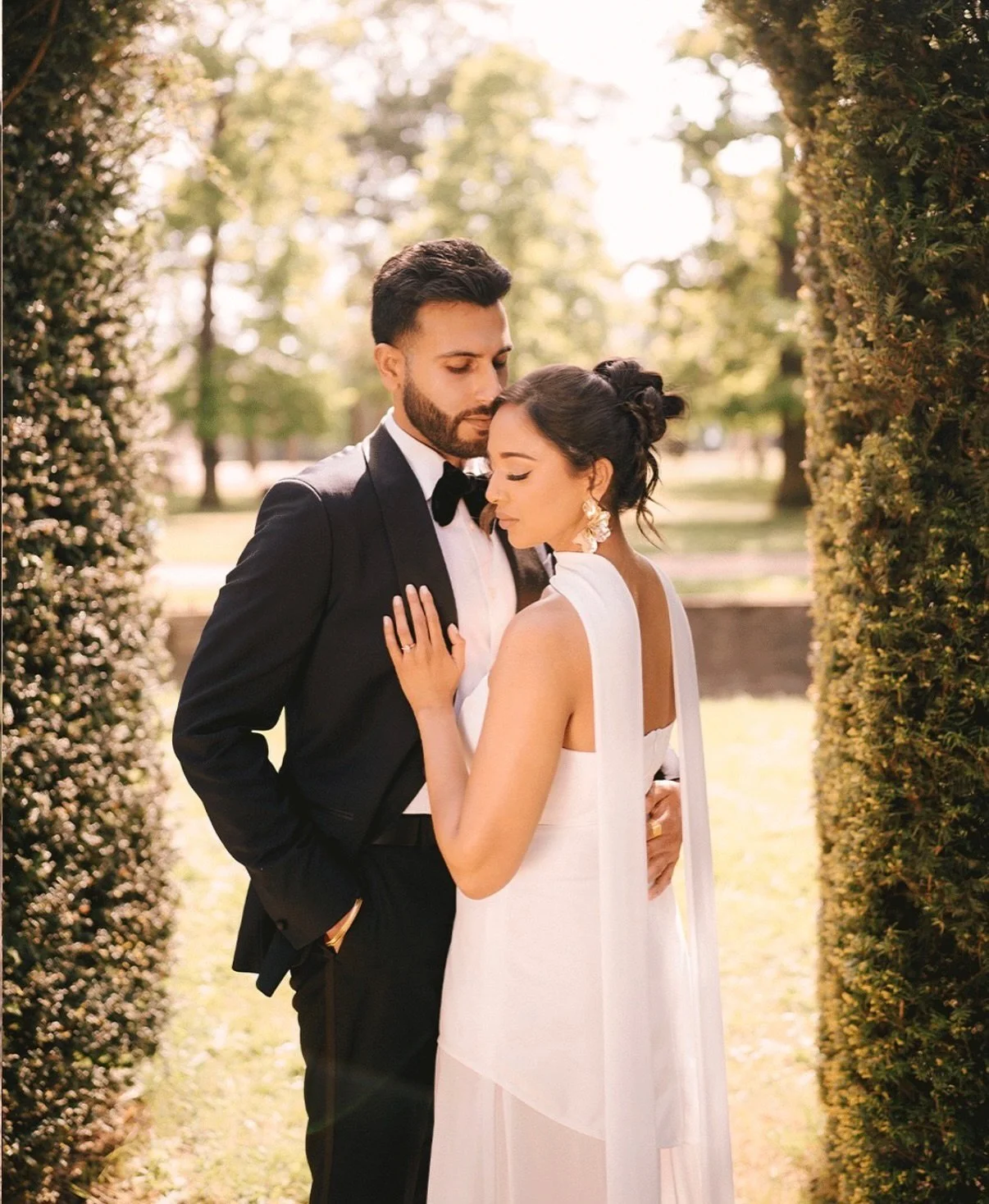 A couple dressed in wedding attire standing close in a sunlit outdoor setting, surrounded by greenery, sharing an intimate moment.