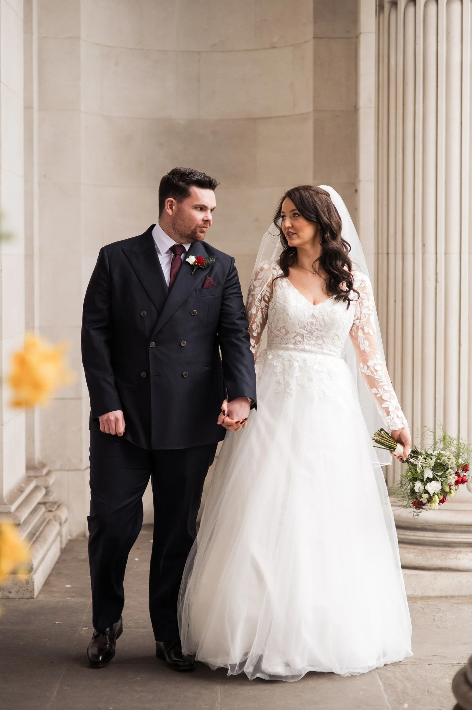 A bride and groom walking together outside a building with columns, holding hands, dressed in wedding attire, and looking at each other.