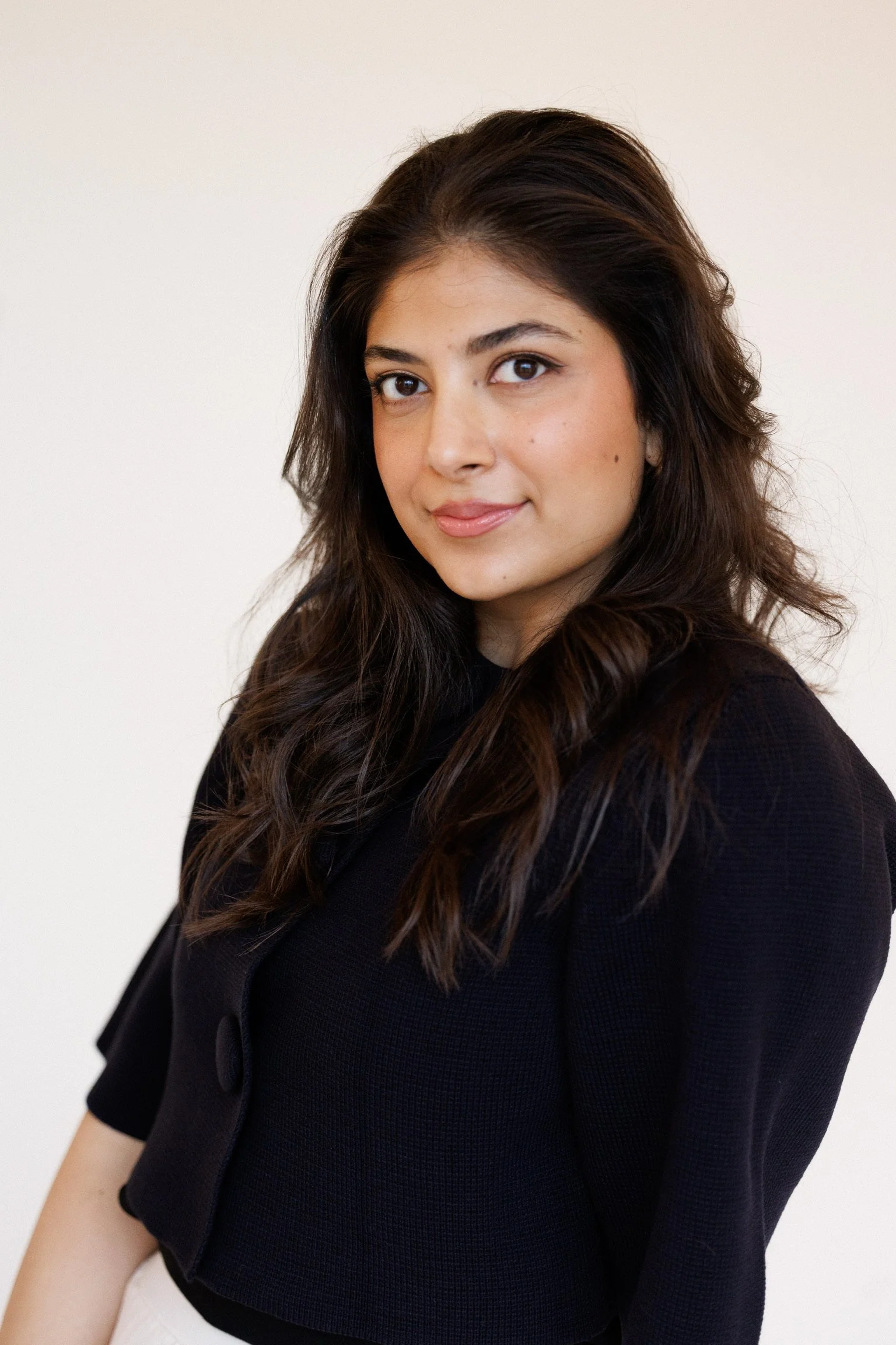 A woman with long, dark, wavy hair and light skin, wearing a black top, is smiling slightly and looking at the camera against a plain white background.