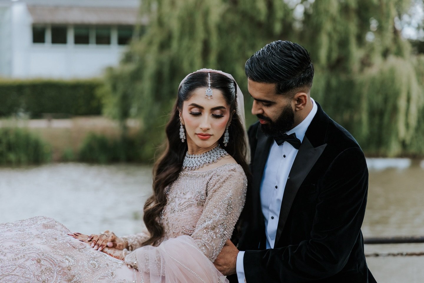 A bride and groom in wedding attire sitting close by a river with trees in the background, dressed elegantly for their wedding.