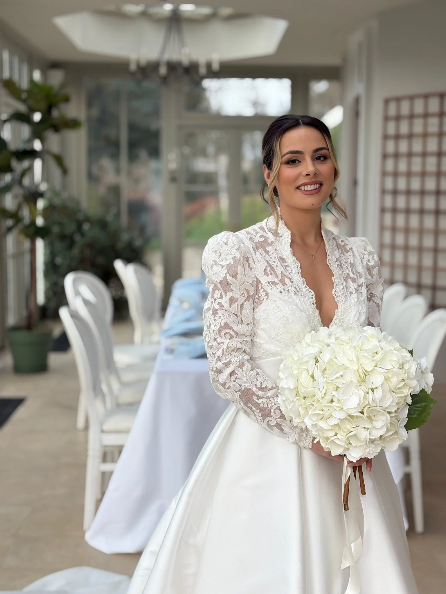 A smiling bride in a lace wedding dress holding a large bouquet of white flowers inside a bright room with white chairs and tables.