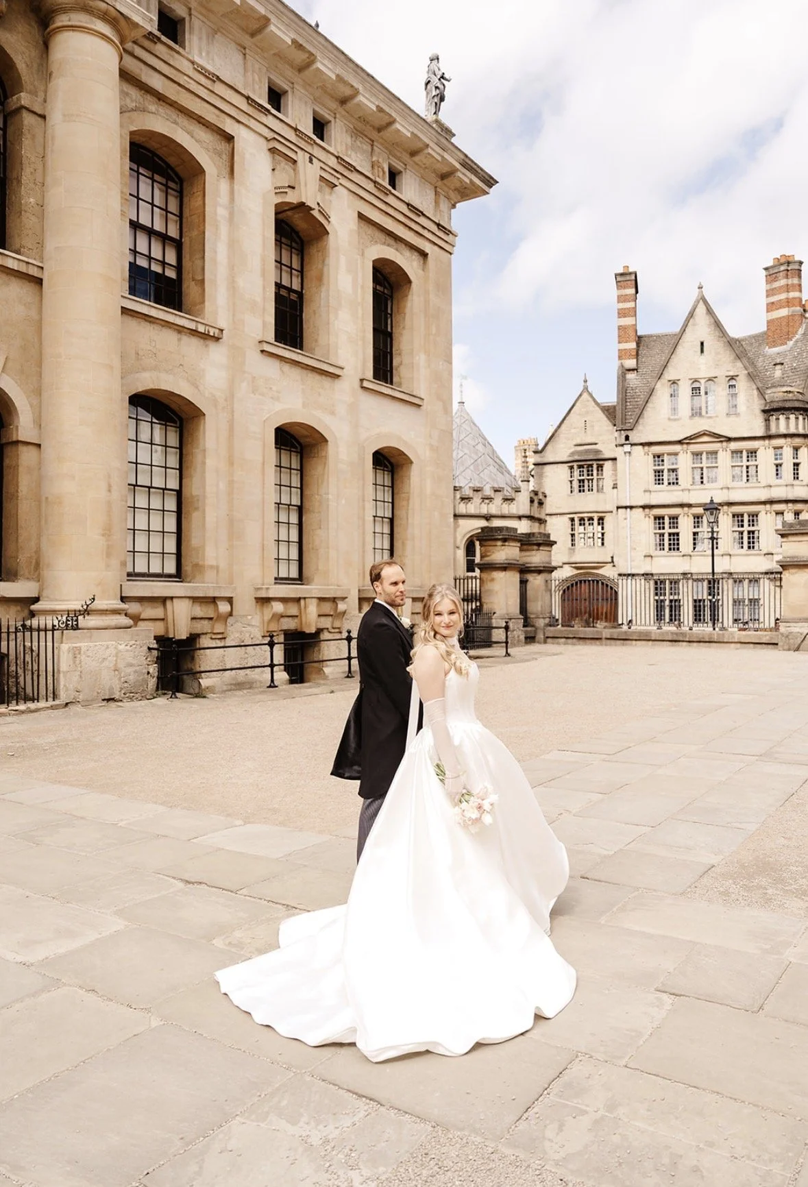 A bride and groom standing outside a historic stone building with large windows. The bride is wearing a white wedding gown and holding a bouquet, while the groom is dressed in a black coat and gray trousers. The sky is partly cloudy.