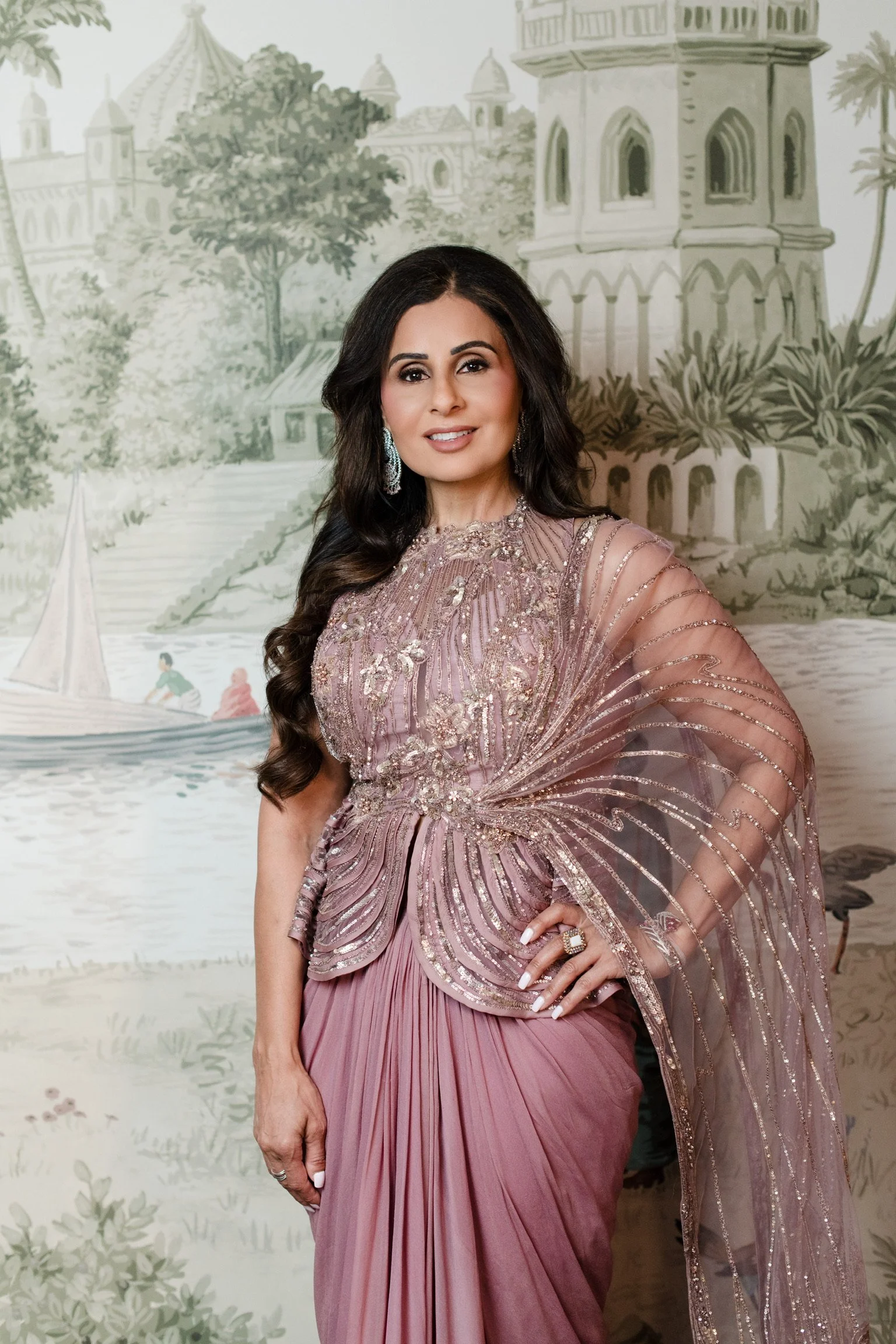 A woman standing in front of a painted backdrop depicting a European-inspired landscape with classical architecture, trees, and a sailboat on the water. She is wearing a pink gown with intricate beading and embroidery, and has long, dark, wavy hair.