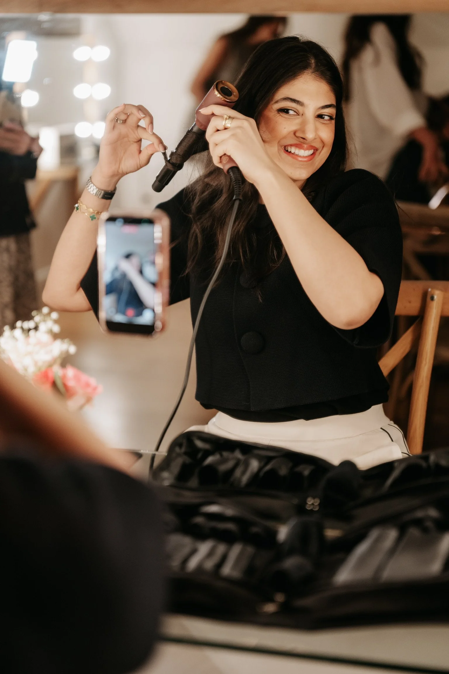 A woman is sitting and smiling as she uses a curling iron to style her hair in a hair salon. She is wearing a black top and white pants, with various accessories including rings, a bracelet, and a watch. The background shows a mirror with lights and other people, likely salon staff or clients.