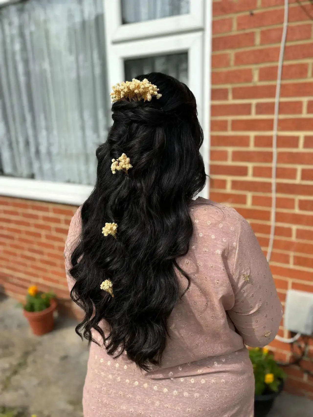 A woman with long, wavy black hair decorated with small white flowers, seen from behind, standing outside in front of a brick wall.