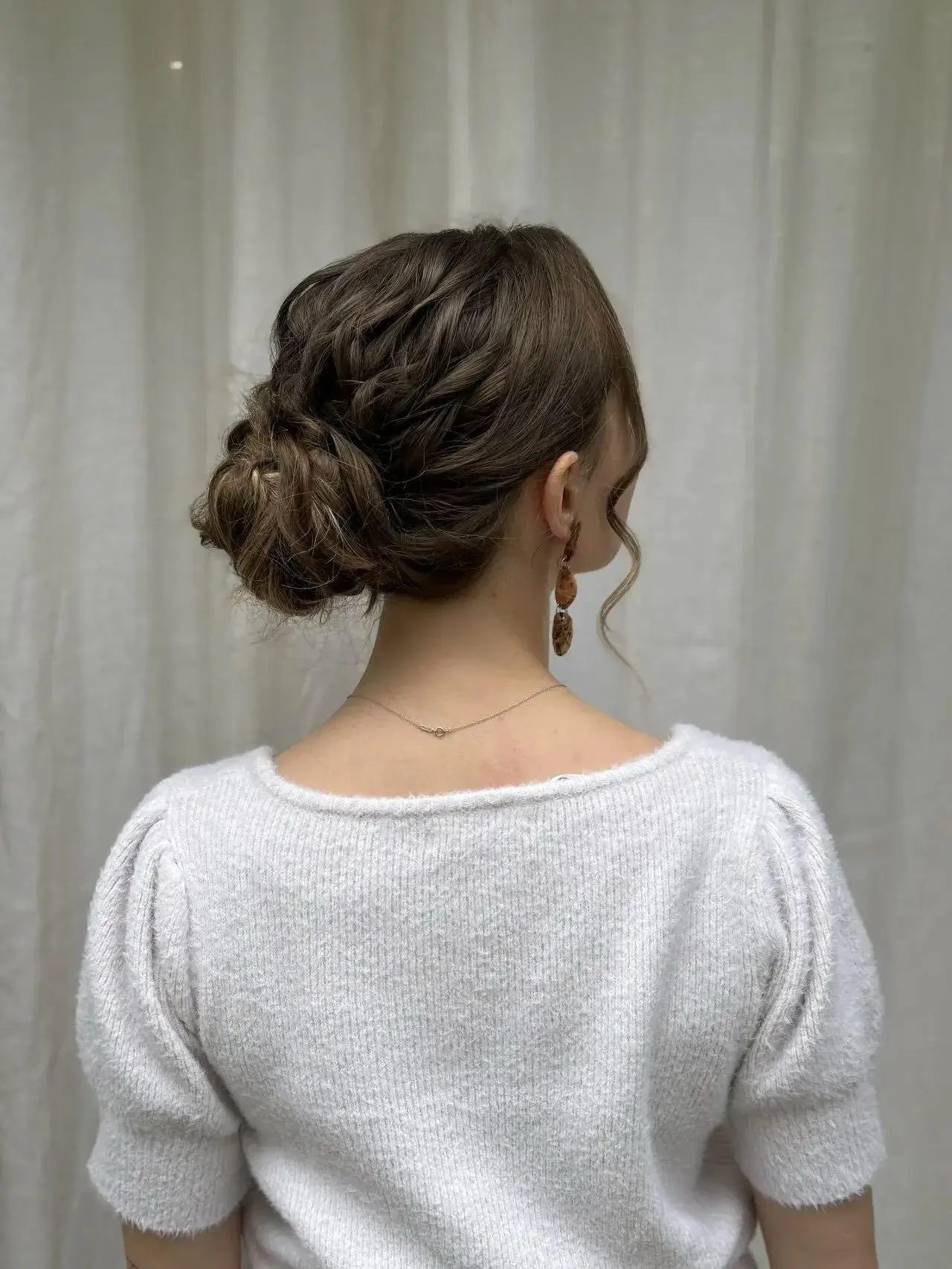 Woman with brown hair styled in a loose, braided updo, wearing a white sweater with puffed sleeves, a delicate necklace, and earrings, standing against a light-colored curtain background.