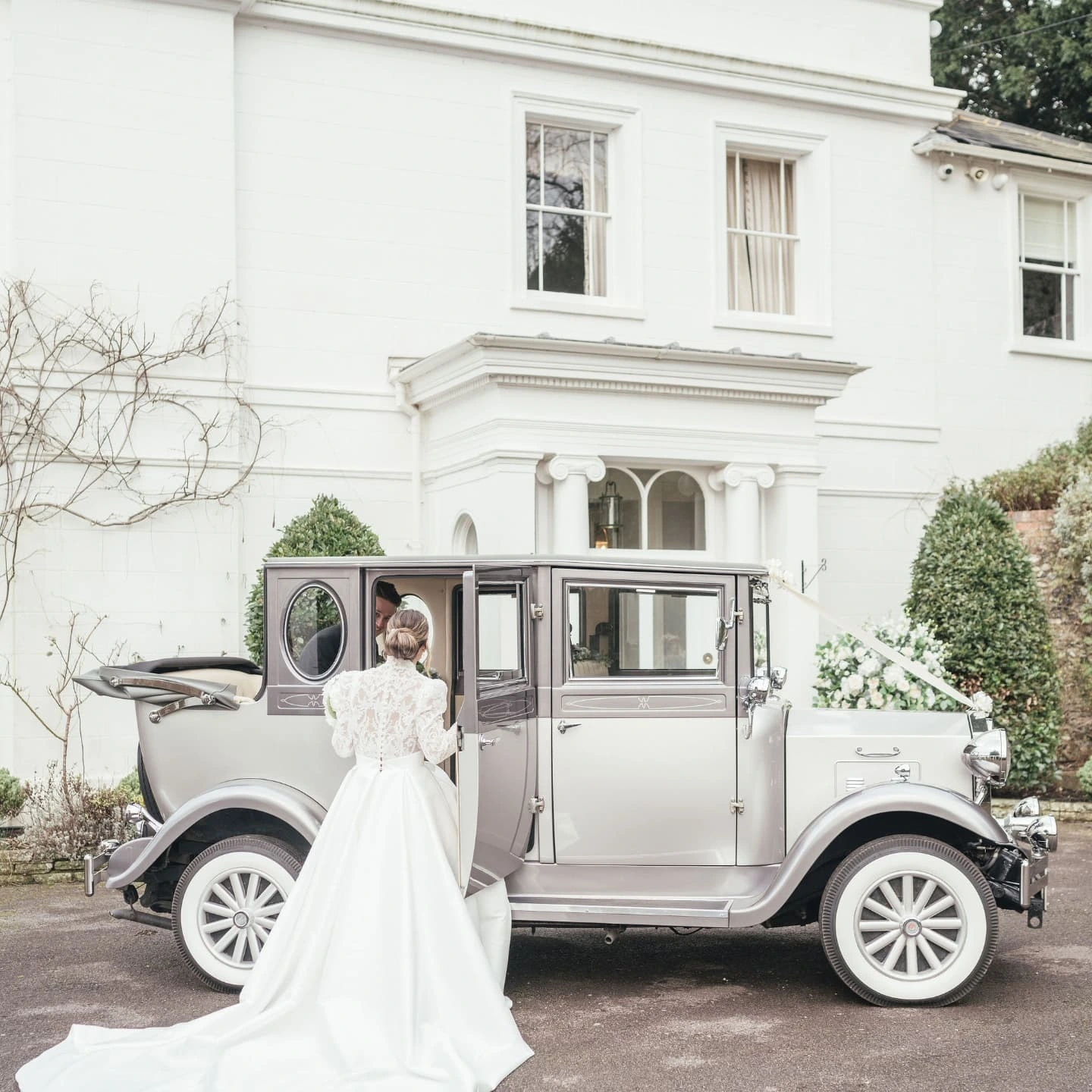 Bride in a white wedding dress standing next to a vintage gray and white car, talking to a person inside the car, in front of a white house with shrubbery.
