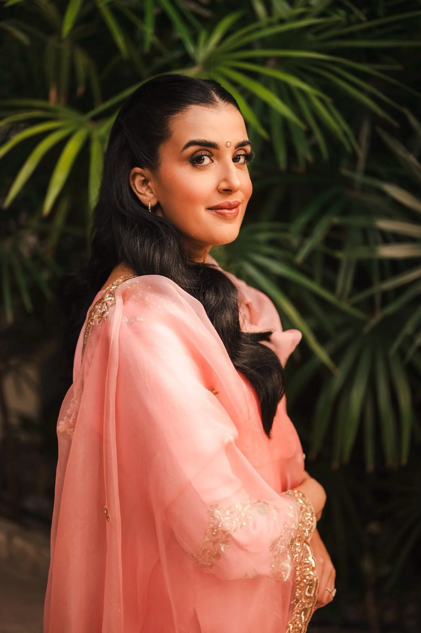 A woman with long dark hair and medium skin tone wearing traditional Indian attire, a pink saree with gold embroidery, standing in front of green leafy plants.