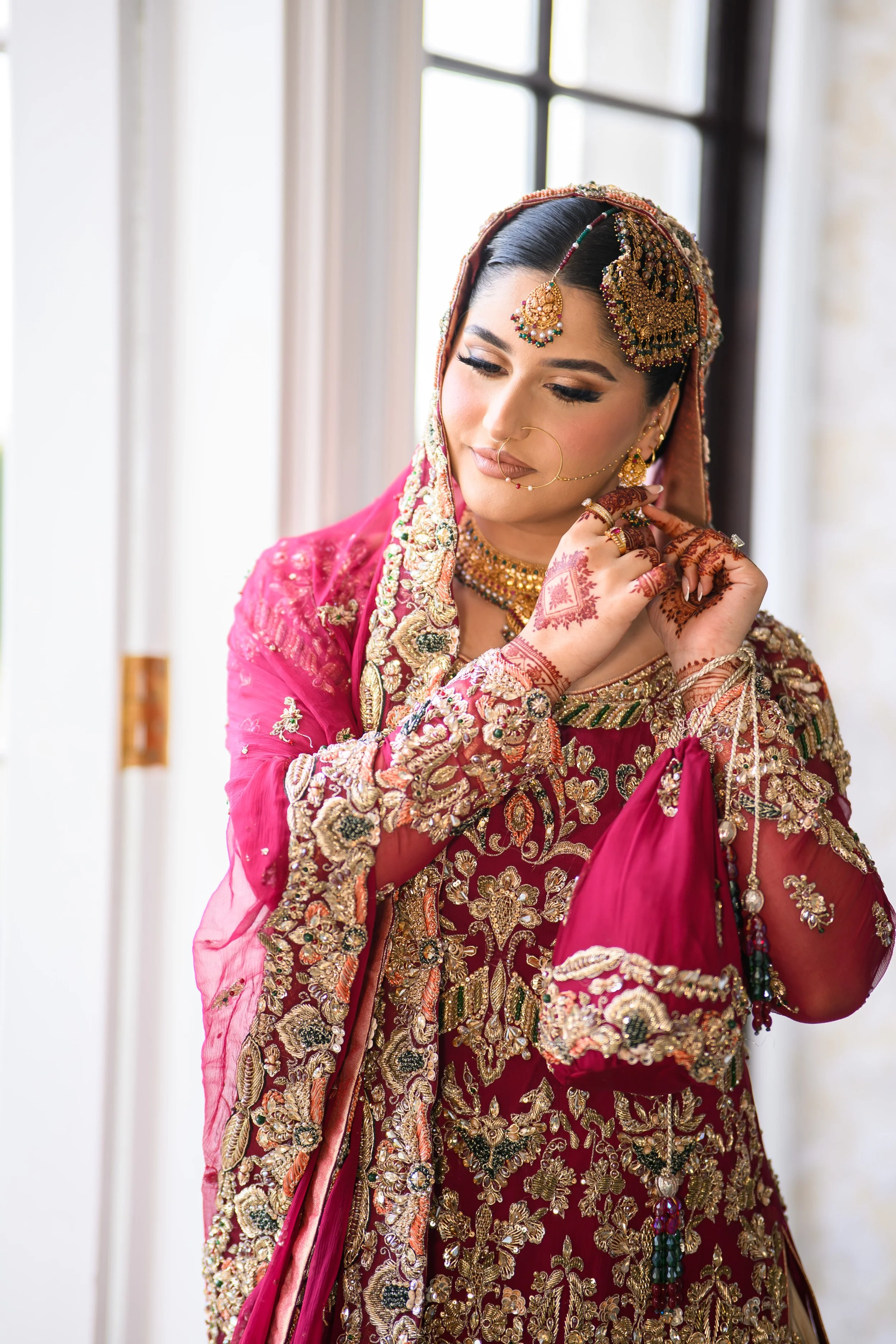 A woman dressed in an ornate pink and gold traditional Indian bridal outfit, wearing elaborate jewelry, including a maang tikka, nose ring, earrings, necklace, and bangles, with henna on her hands, standing near a window.
